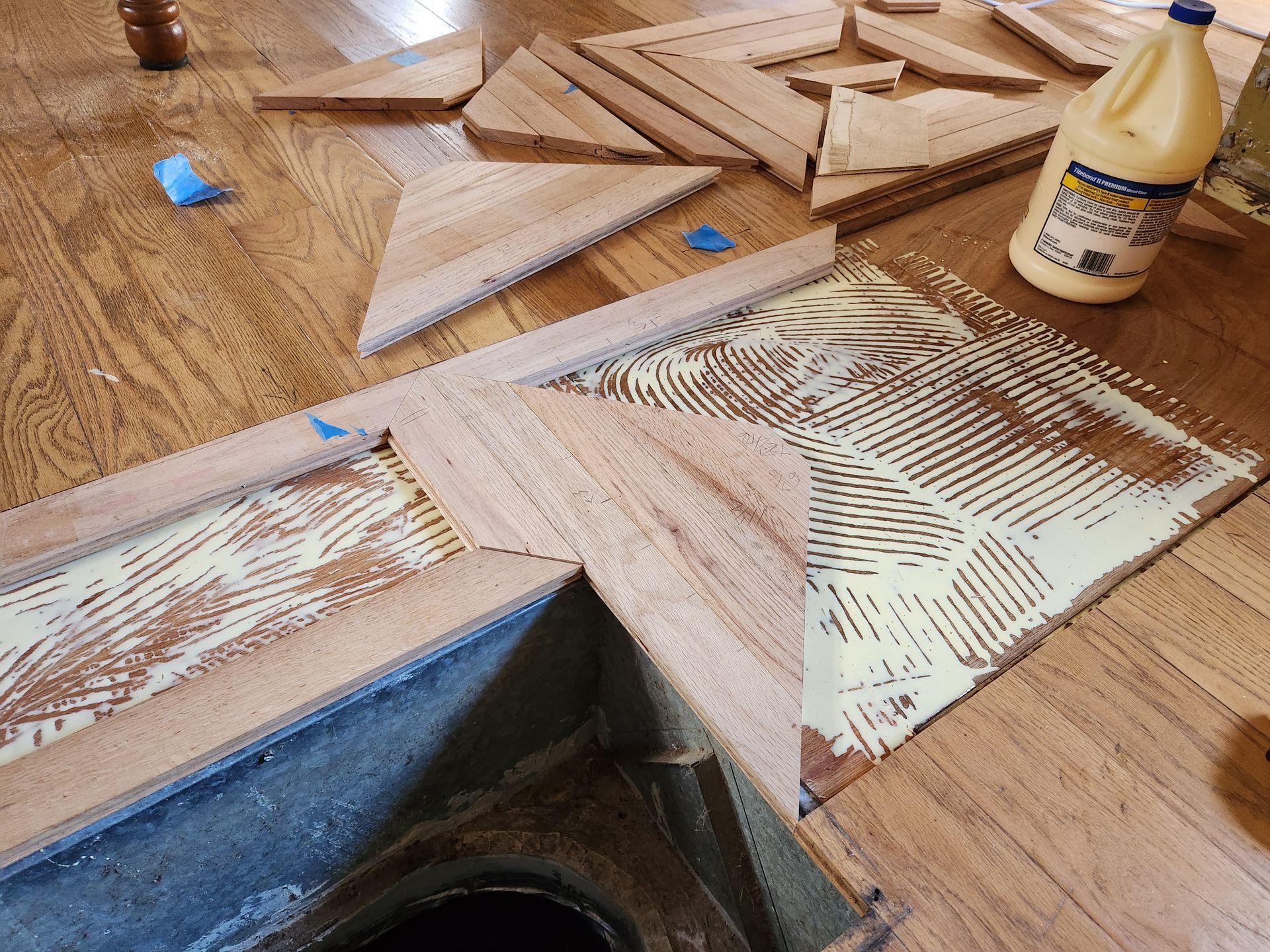 Hardwood floor pieces being installed over adhesive near a floor vent with a bottle of wood glue nearby.