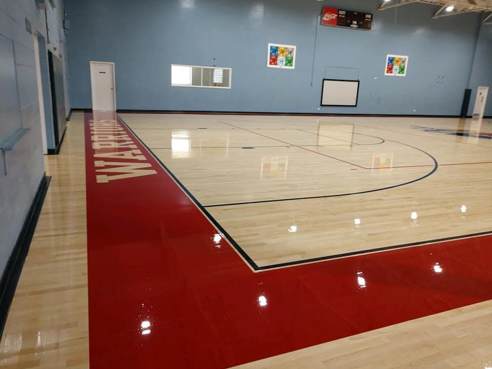 An indoor basketball court with light wood flooring, red painted borders, and the word 