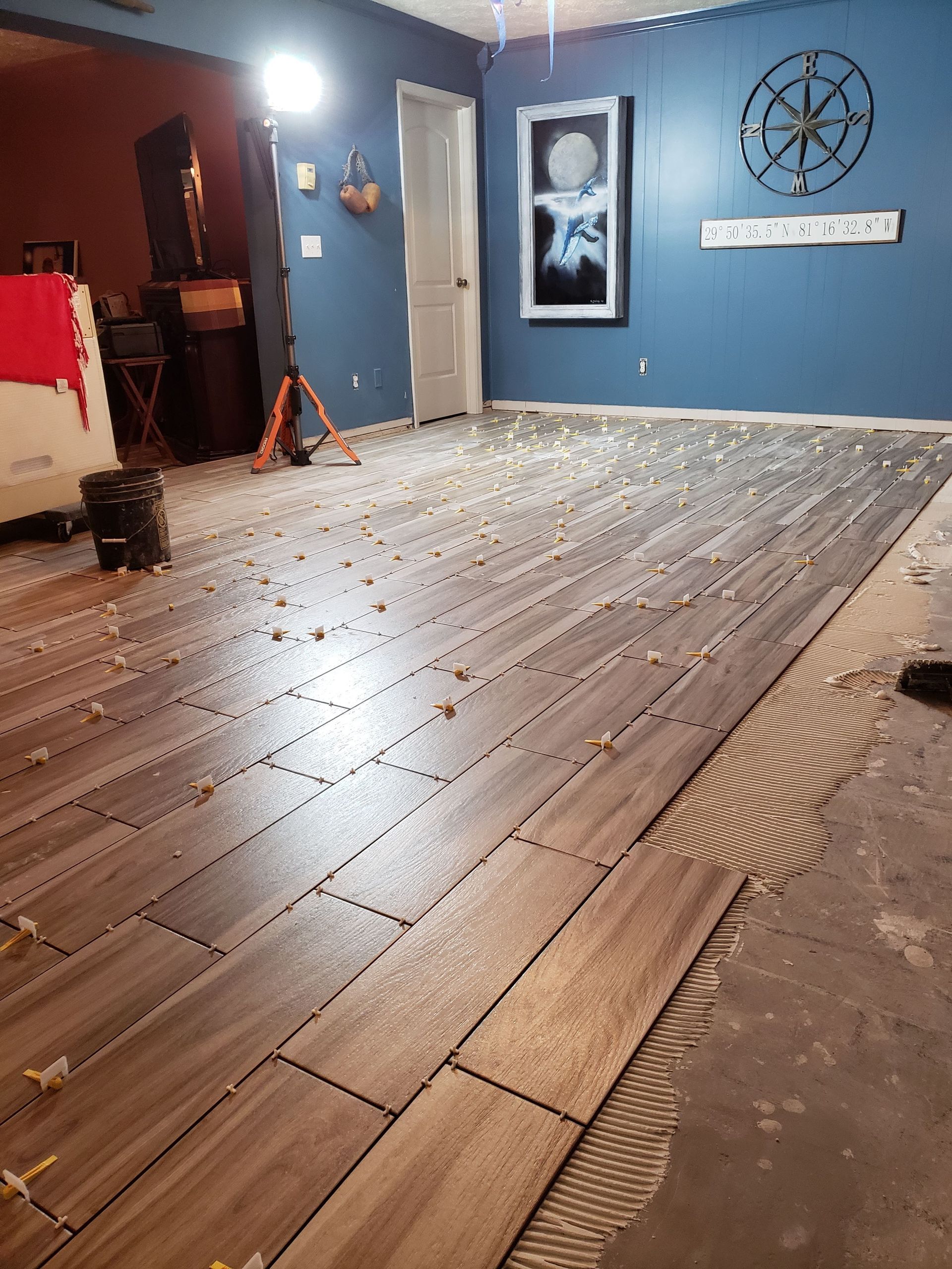 A partially installed wood-look tile floor with leveling spacers on top of combed mortar in a blue-walled room.
