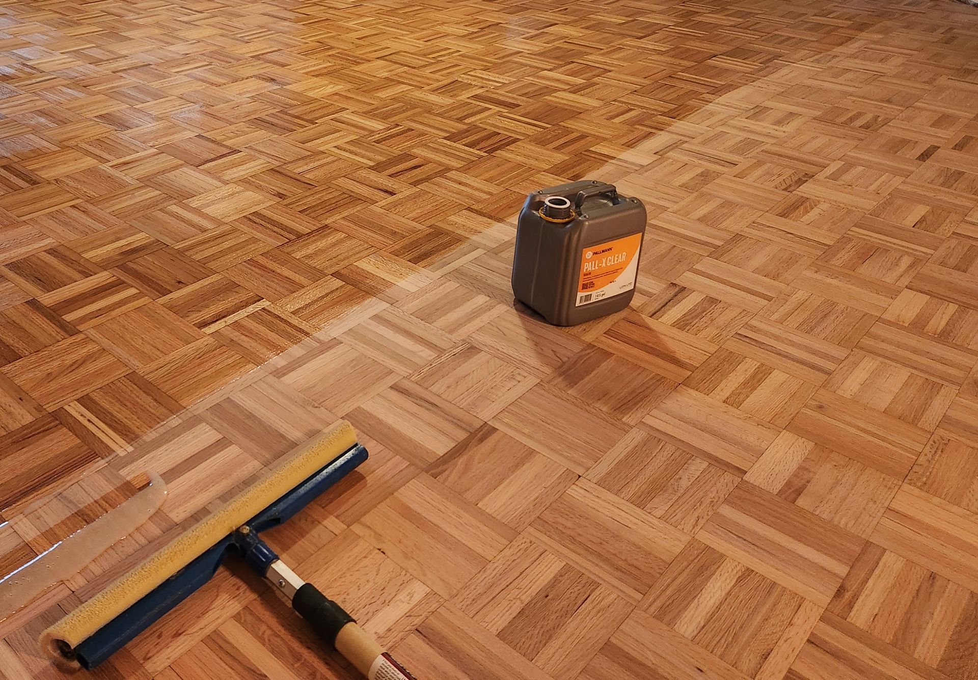 A squeegee and a container of finish sit on a wooden parquet floor, showing the progress of a refinishing project.