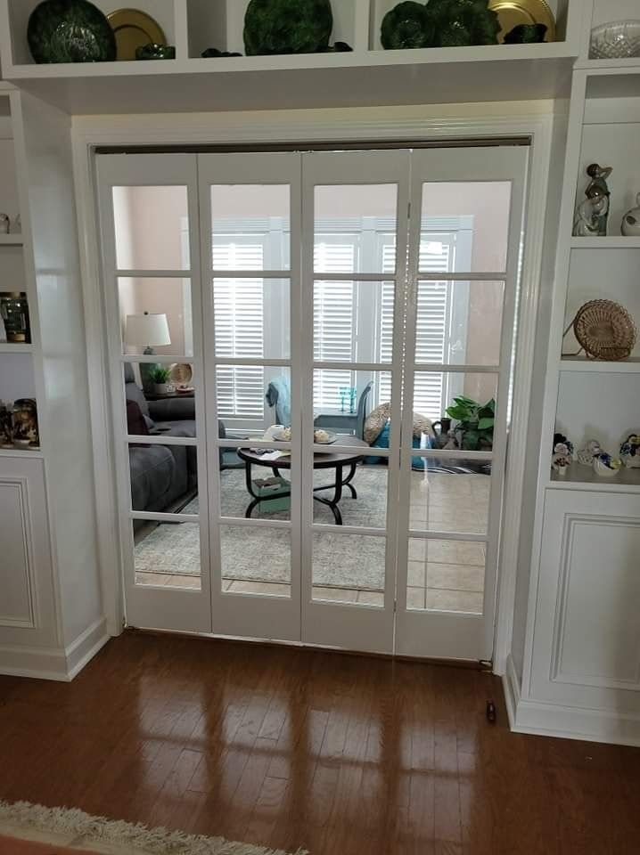 White framed glass doors lead into a living room with a couch, table, and windows with shutters.