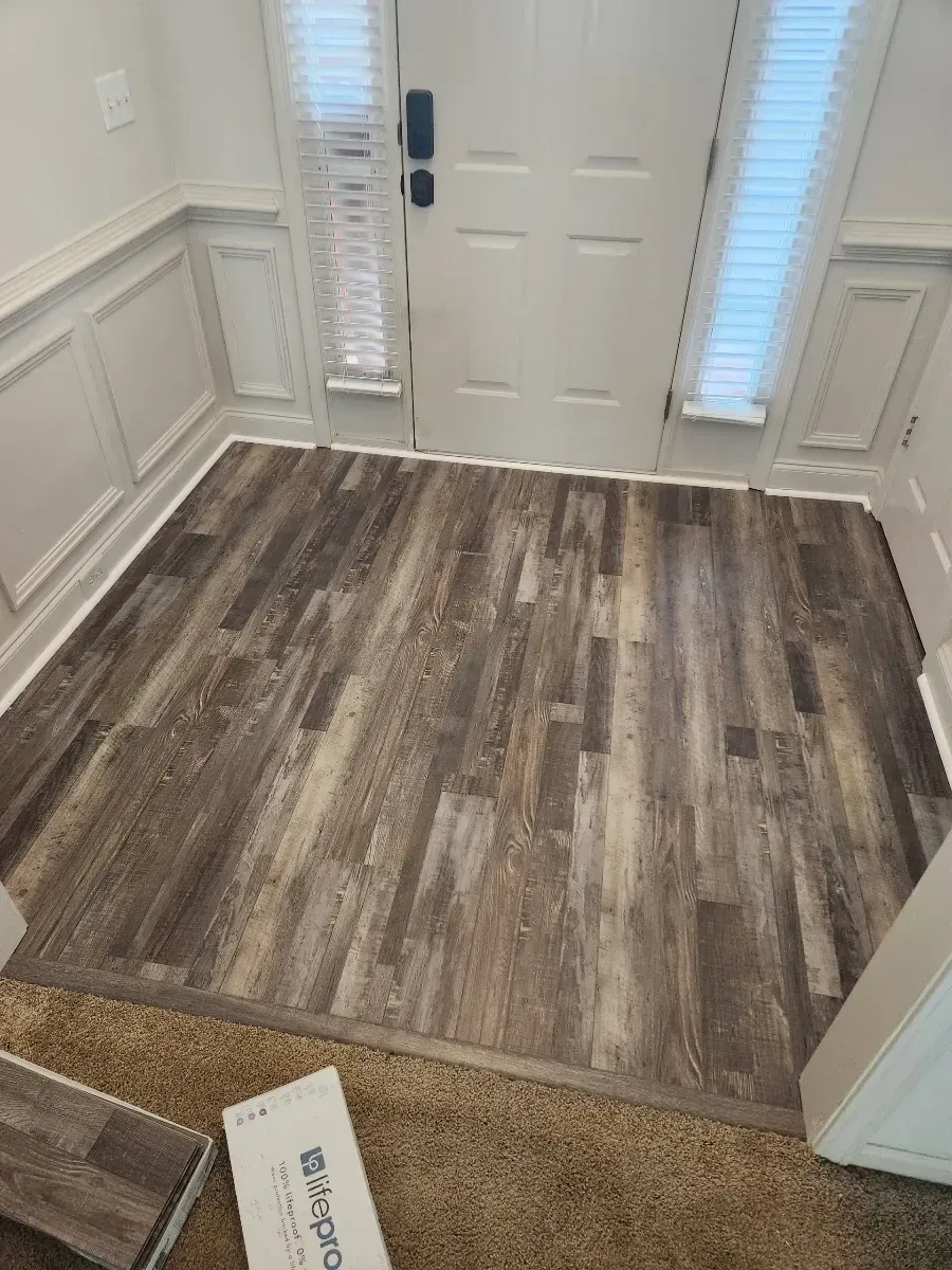 Entryway with wood-look flooring, white door, sidelights, and wainscoting. A sample of the flooring is on the carpet.