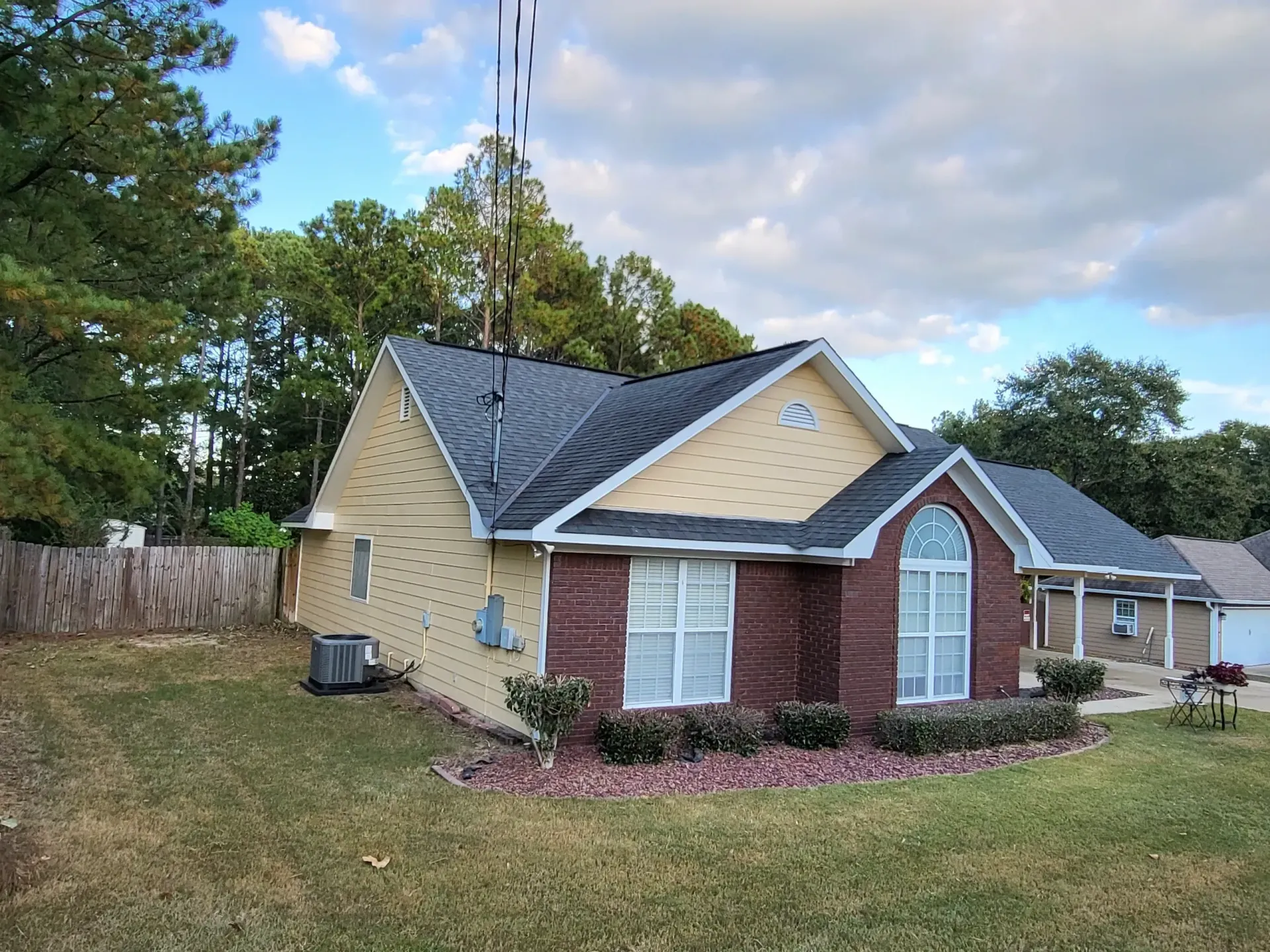 A brick and yellow house with a dark roof, in front of a green lawn and trees, under a cloudy sky.