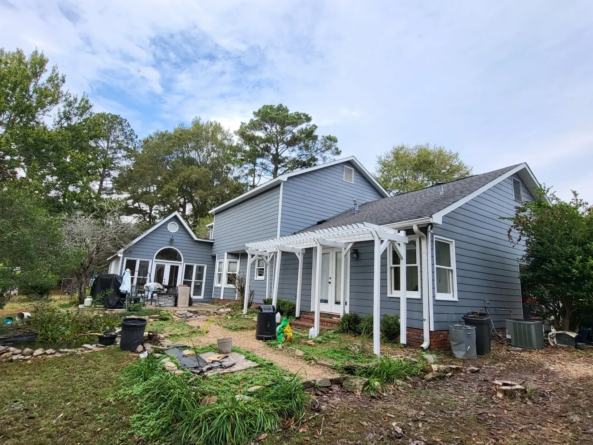 A blue-gray house with white trim and a pergola, surrounded by overgrown landscaping under a cloudy sky.