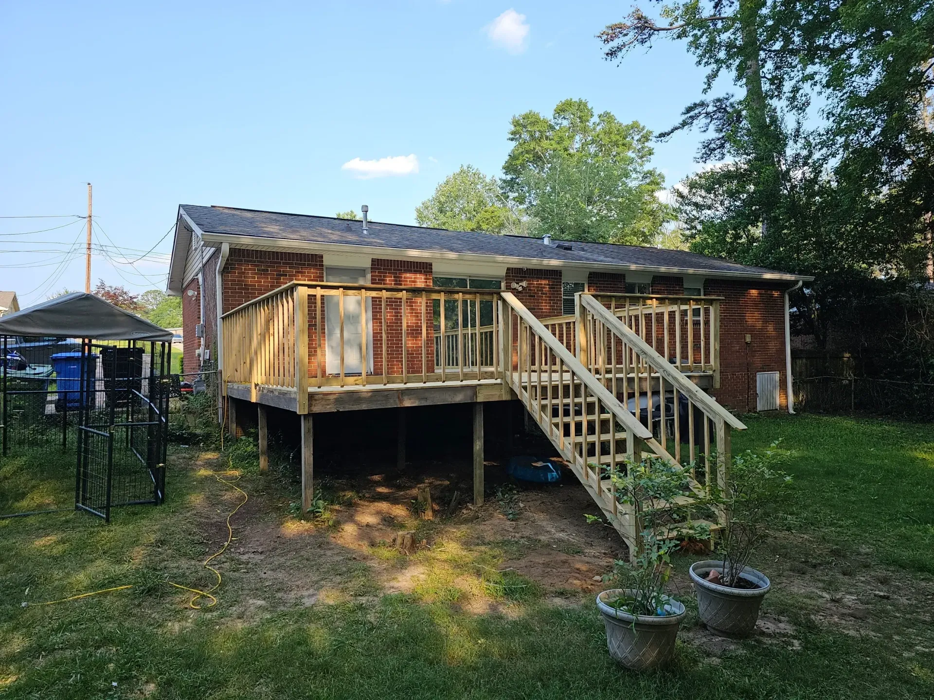 Brick house with wooden deck and stairs in a grassy backyard, on a sunny day.