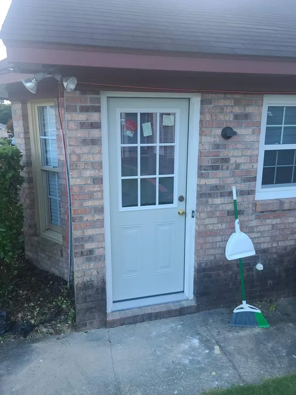 Brick building with white door and windows; broom and dustpan rest beside the door.
