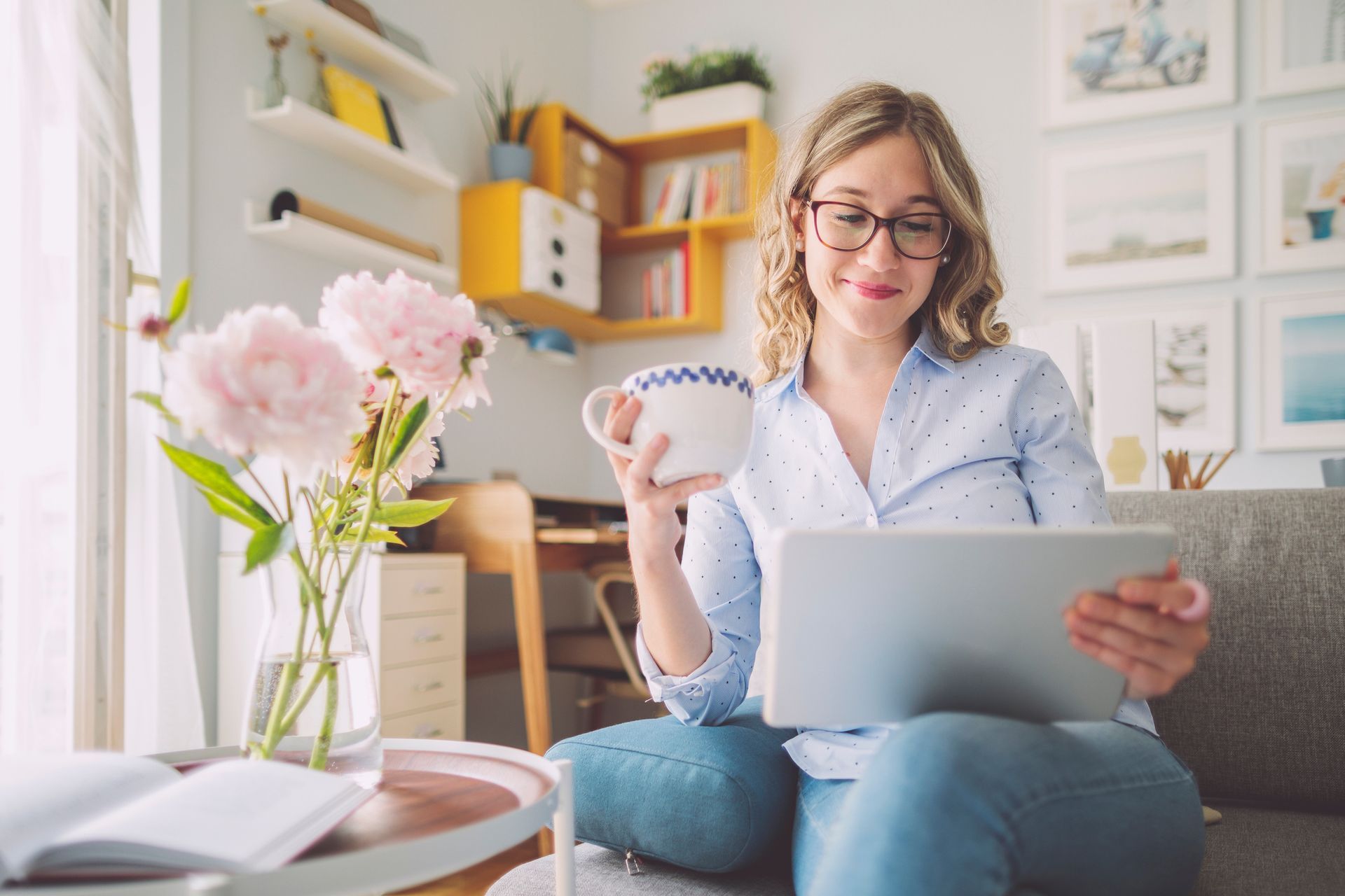 A woman is sitting on a couch holding a cup of coffee and a tablet.