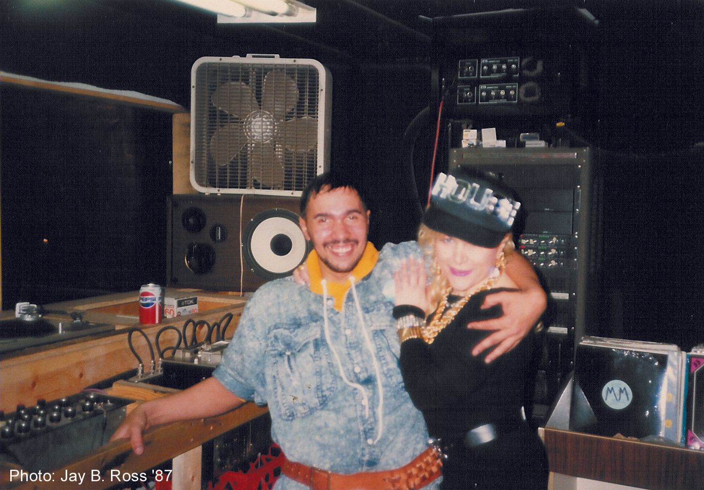 A man and a woman are posing for a photo in a room with a fan and speakers