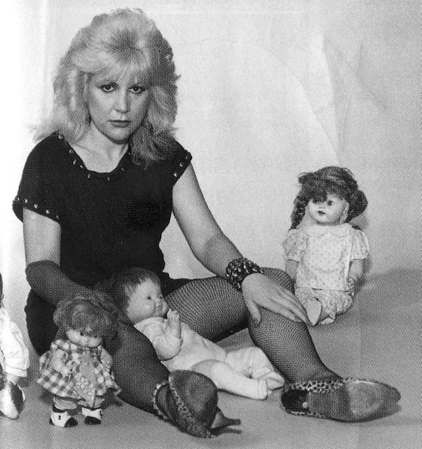 A woman sits on the floor surrounded by dolls