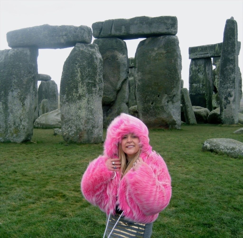 A woman in a pink fur coat stands in front of stone structures