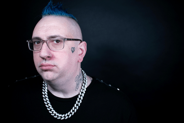 A man with blue hair and glasses is wearing a chain around his neck.