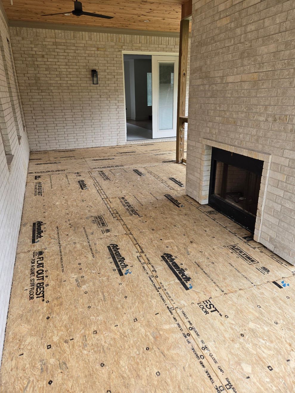 Unfinished room with exposed brick walls, plywood subfloor, and a fireplace opening near French doors. Unfinished room with exposed brick walls, plywood subfloor, and a fireplace opening near French doors.