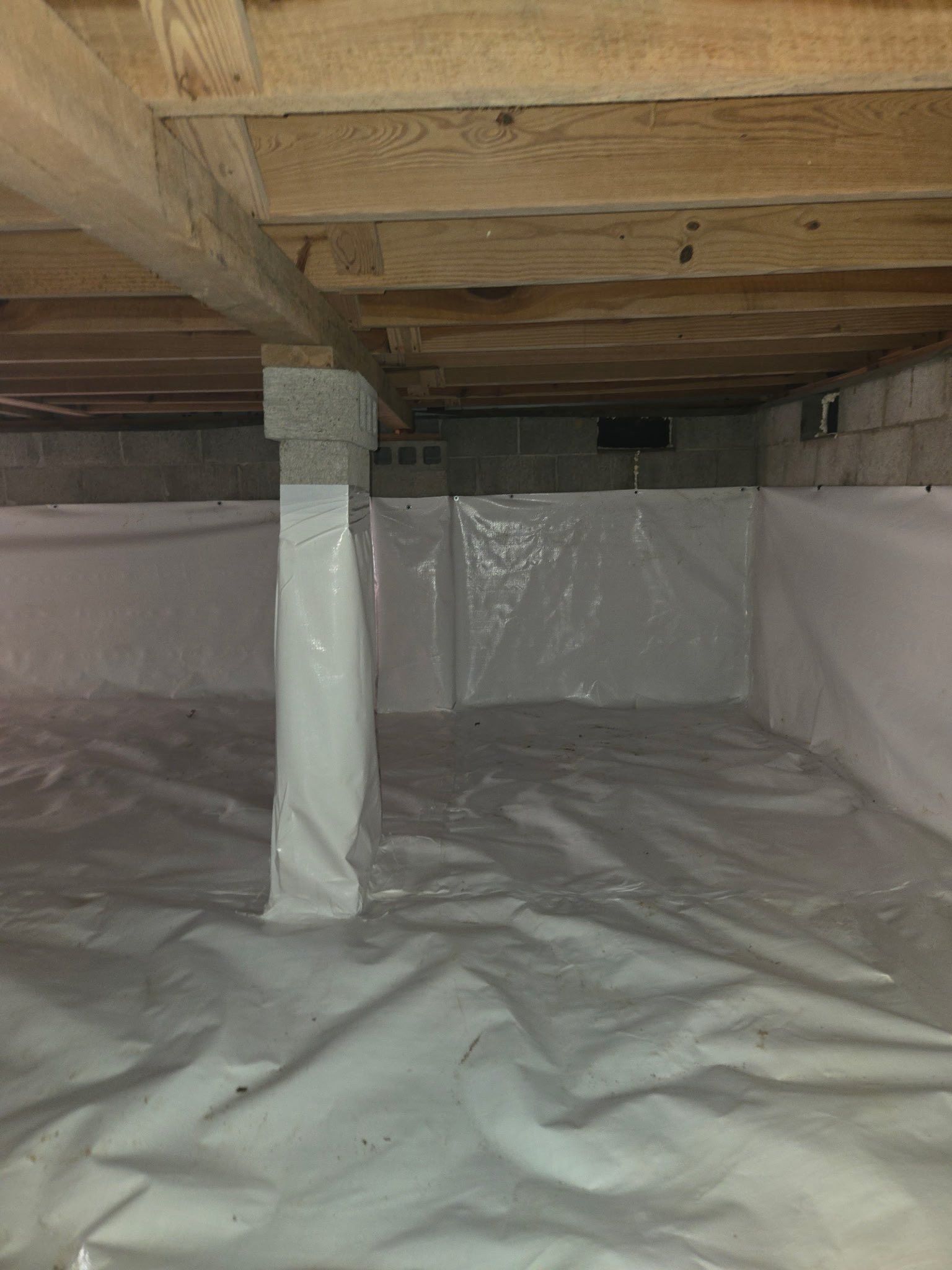 Crawlspace with exposed wooden beams, wrapped support column, and plastic vapor barrier on the floor and walls