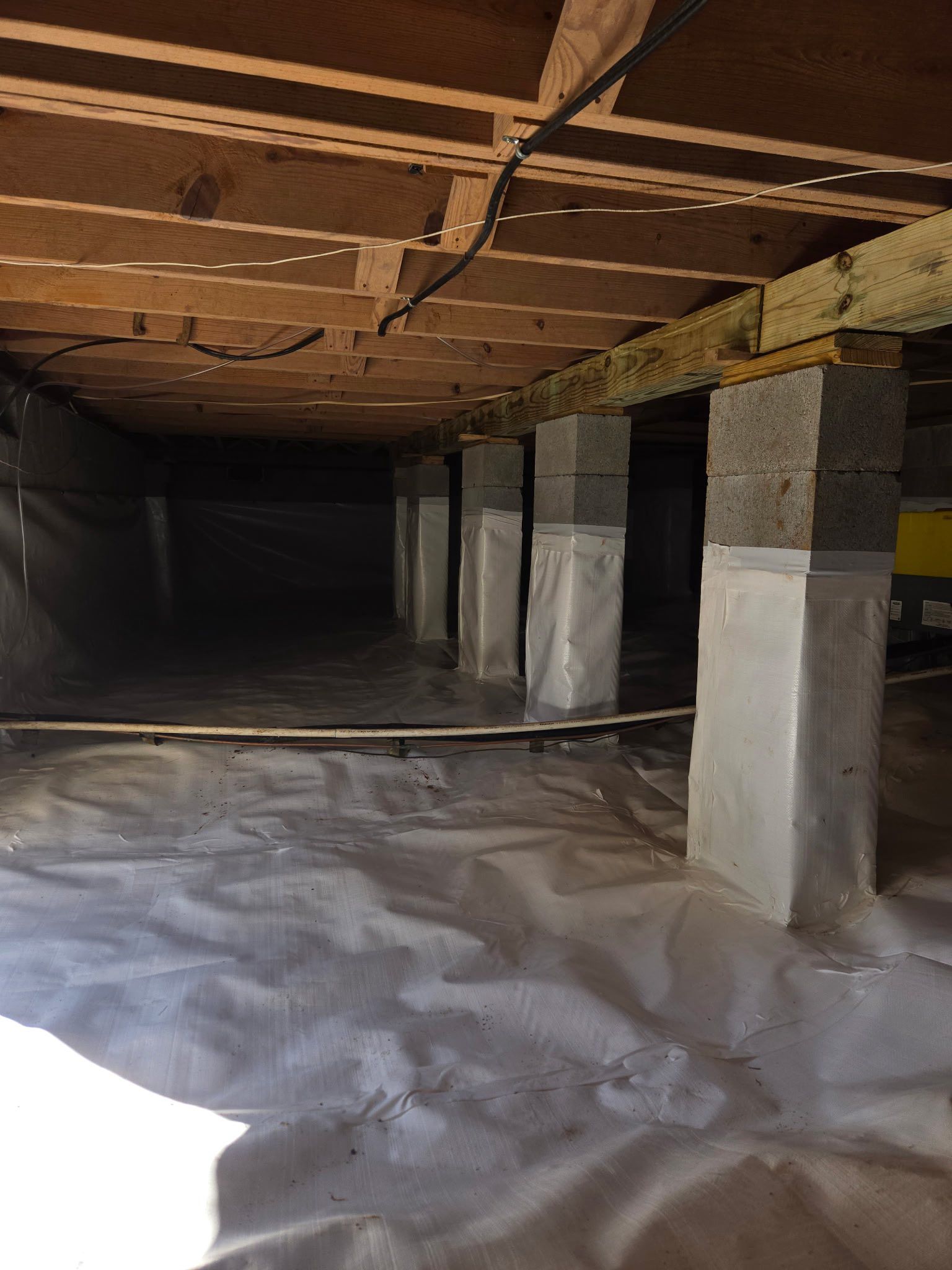 Crawlspace under a house with exposed beams, concrete supports, and plastic sheeting on the ground.