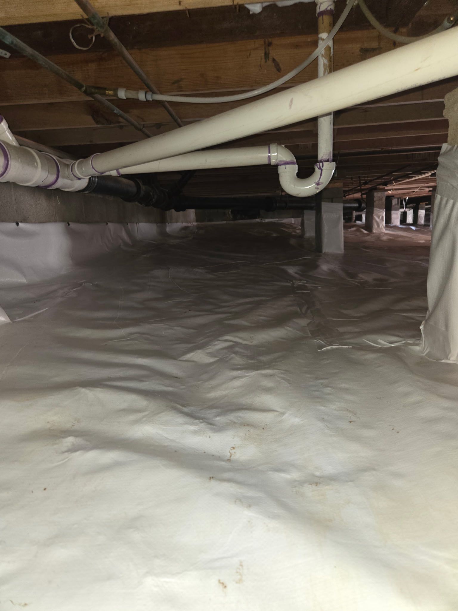 Crawlspace with plastic vapor barrier on the floor and exposed pipes and beams overhead