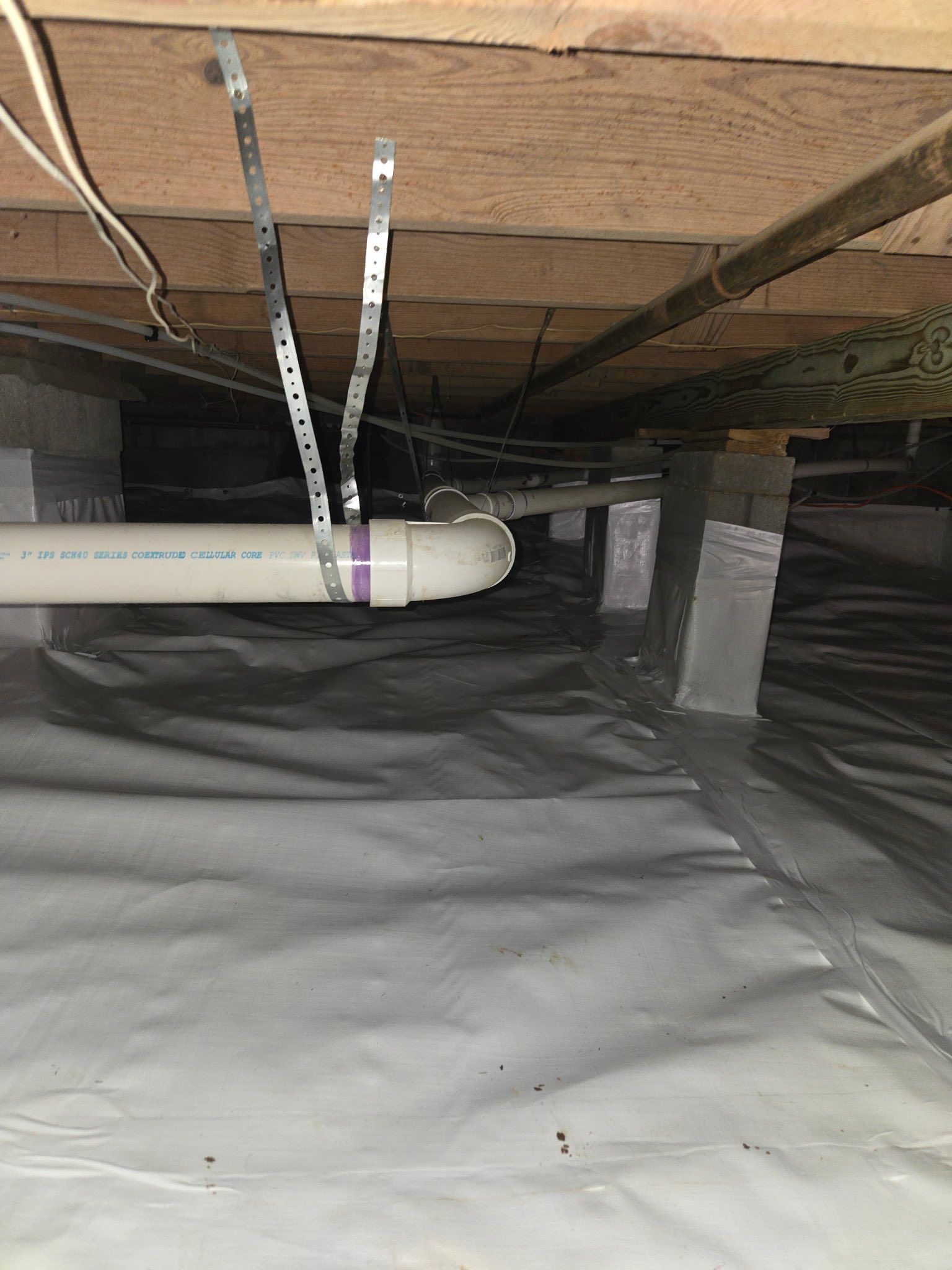 Under-floor crawlspace with white PVC pipe, insulation, and black vapor barrier lining the ground. Under-floor crawlspace with white PVC pipe, insulation, and black vapor barrier lining the ground.