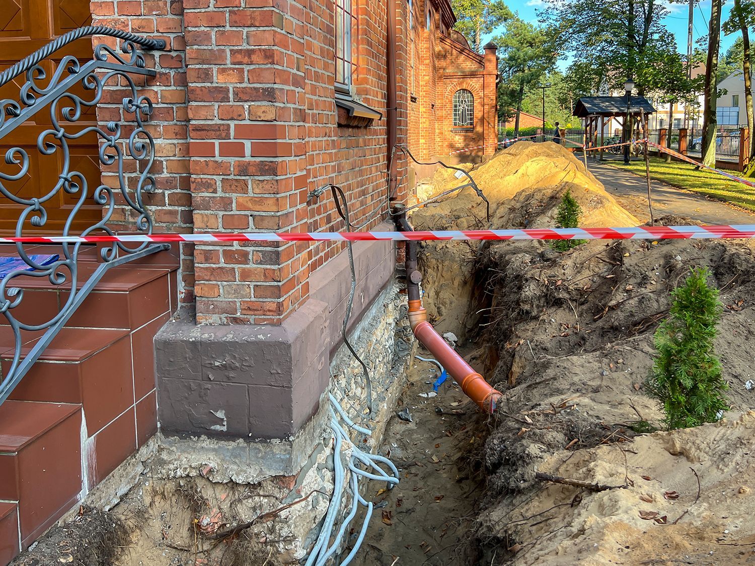 Partially collapsed ground beside brick house, with exposed pipes and caution tape near a trench Partially collapsed ground beside brick house, with exposed pipes and caution tape near a trench
