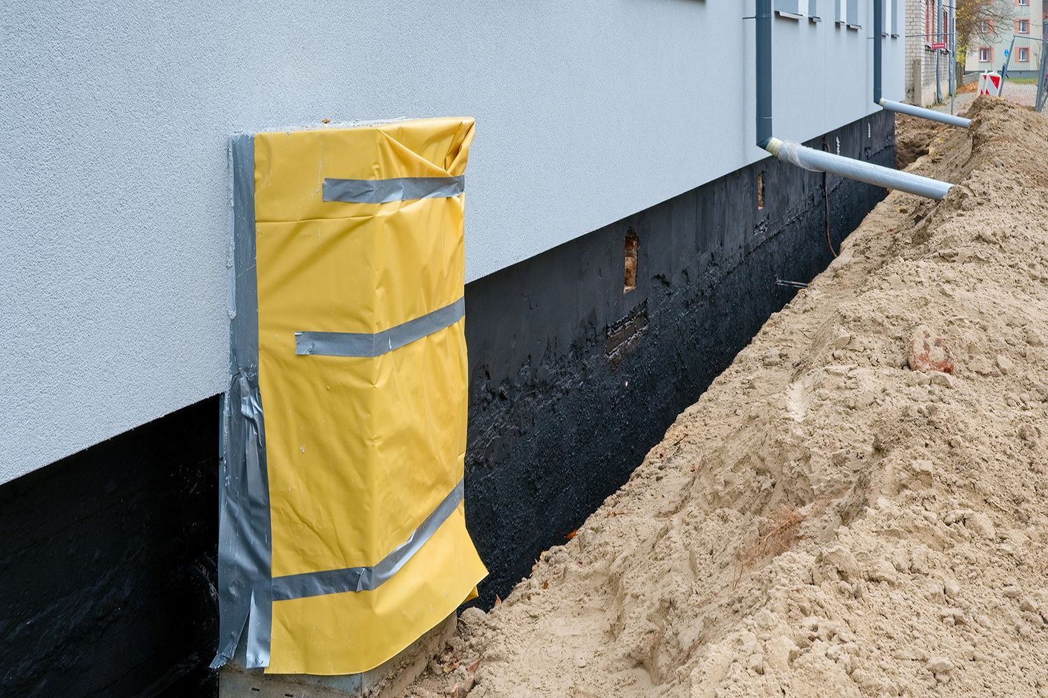Yellow protective wrap on a building corner beside a black waterproofed foundation and sandy trench. Yellow protective wrap on a building corner beside a black waterproofed foundation and sandy trench.