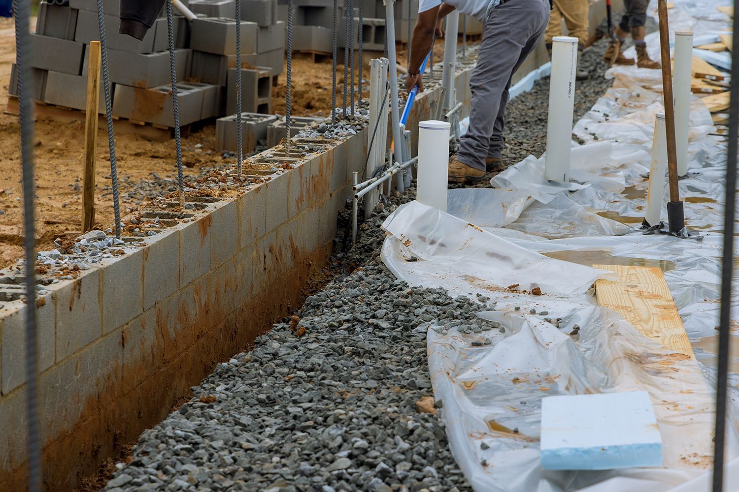 Construction site with gravel path, concrete wall, white plastic sheeting, and workers' legs visible. Construction site with gravel path, concrete wall, white plastic sheeting, and workers' legs visible.