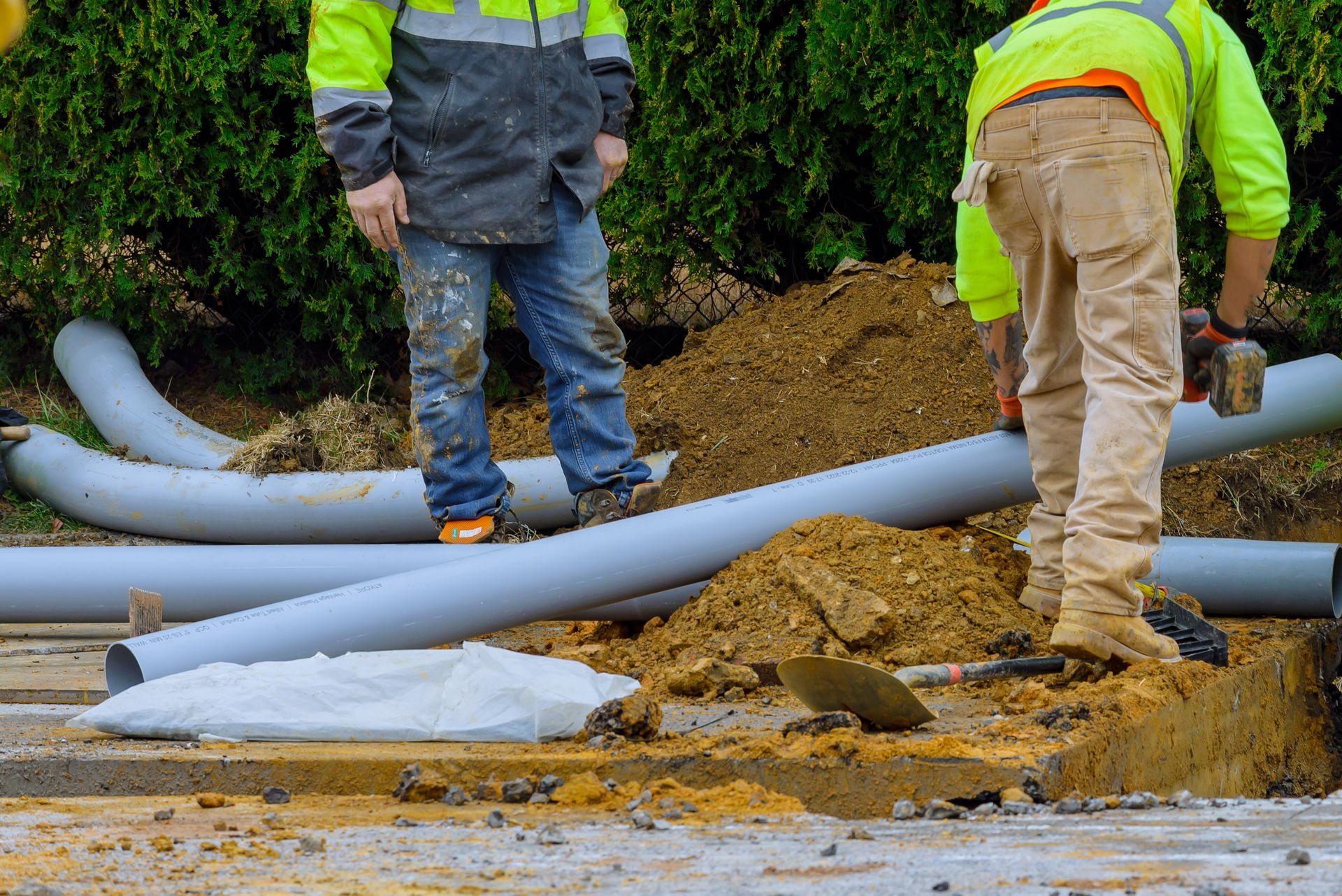 Workers handling gray utility pipes at a muddy roadside construction site