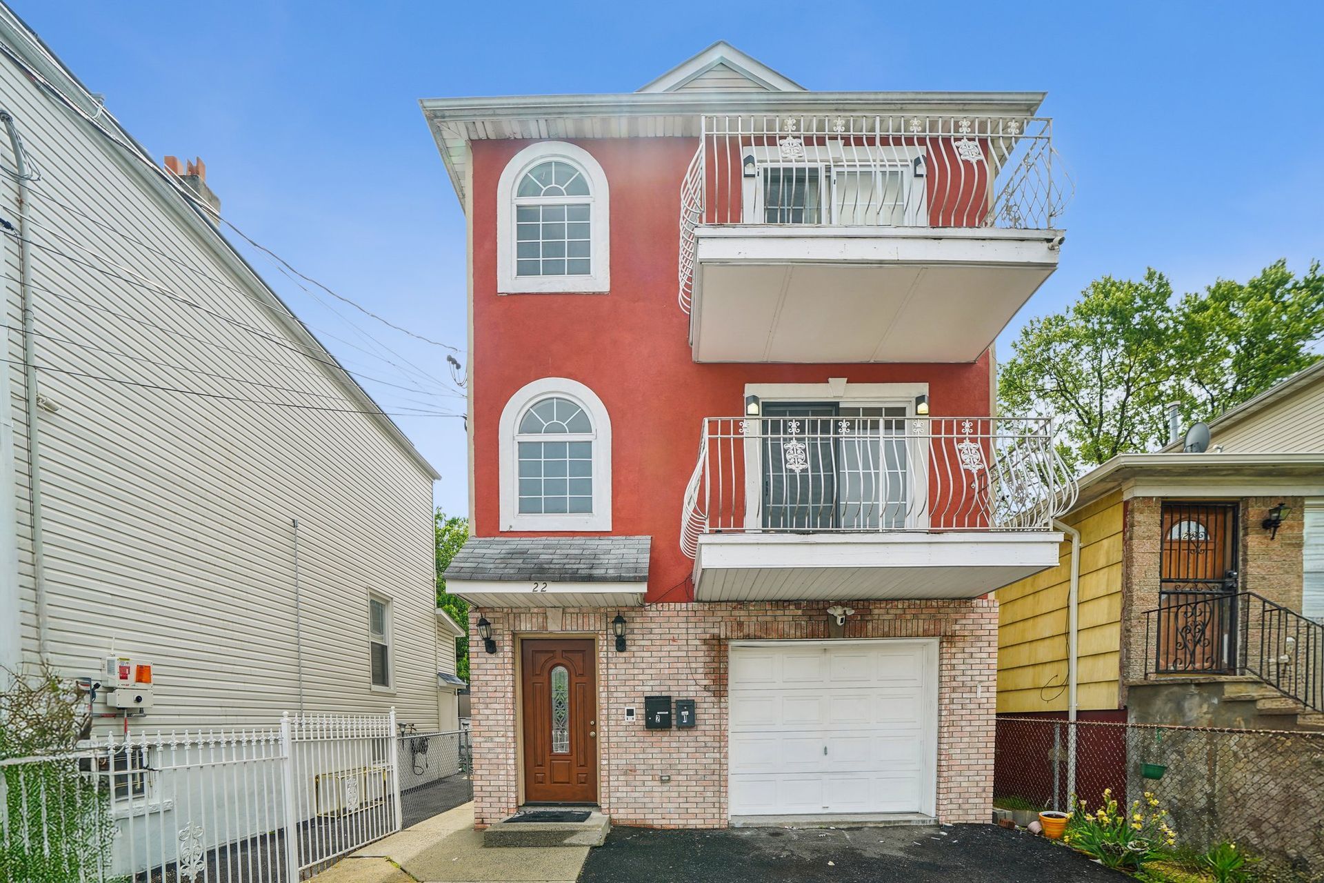 Red stucco multi-story home with balconies, arched windows, and garage door, between neighboring houses.