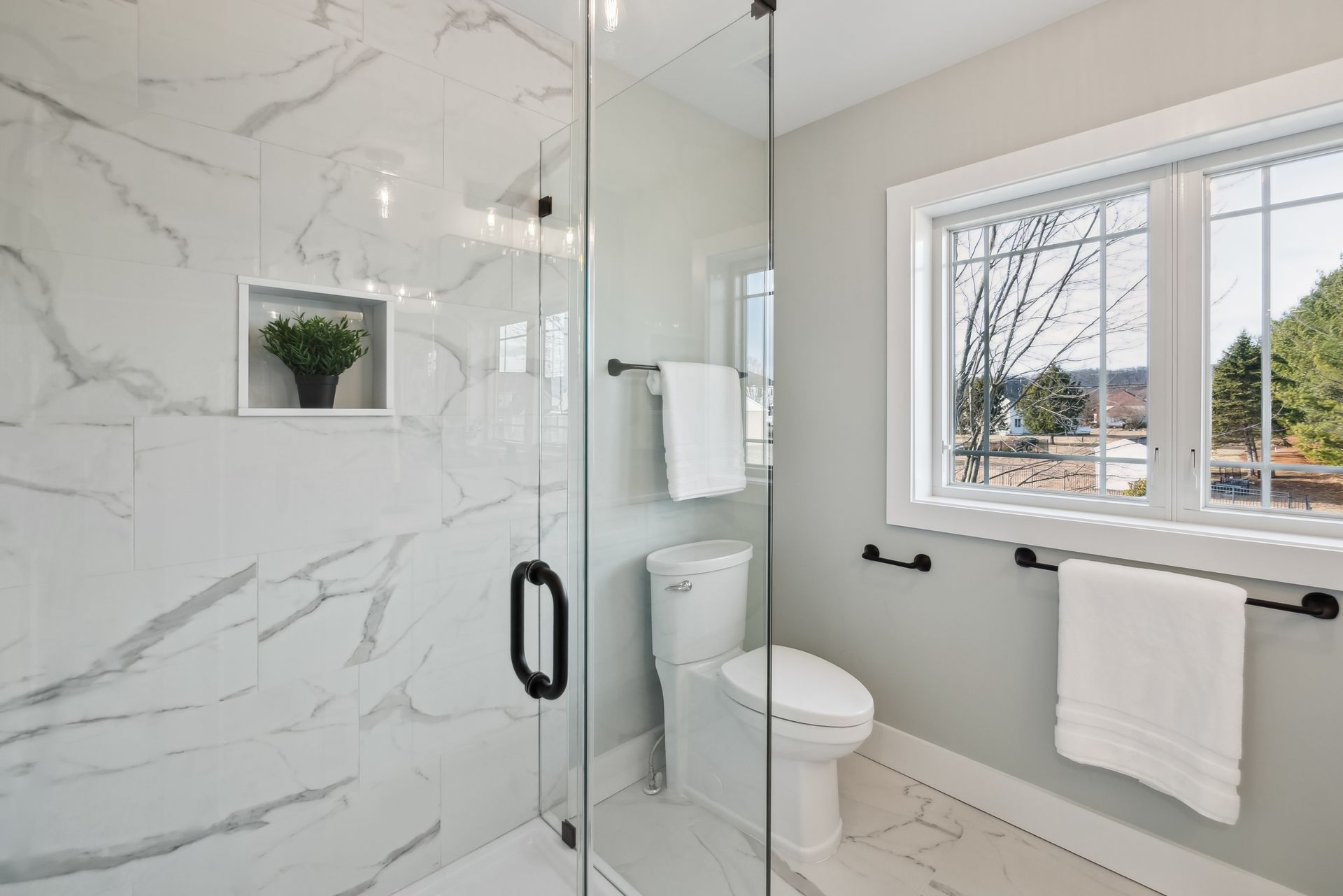 Bathroom with marble shower, toilet, window, and towel racks. White and gray tones.