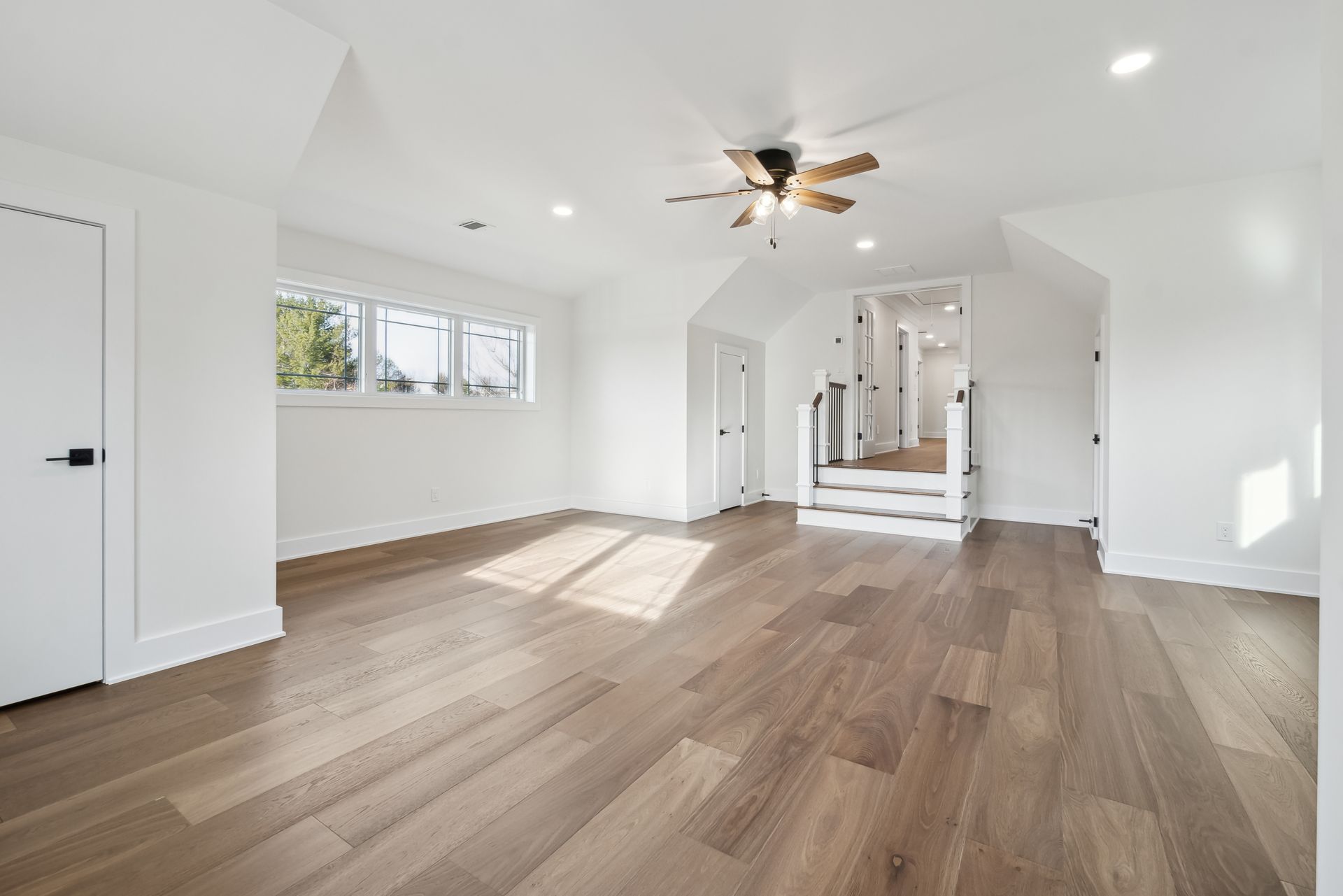 Bright, empty room with hardwood floors, white walls, windows, and a ceiling fan.