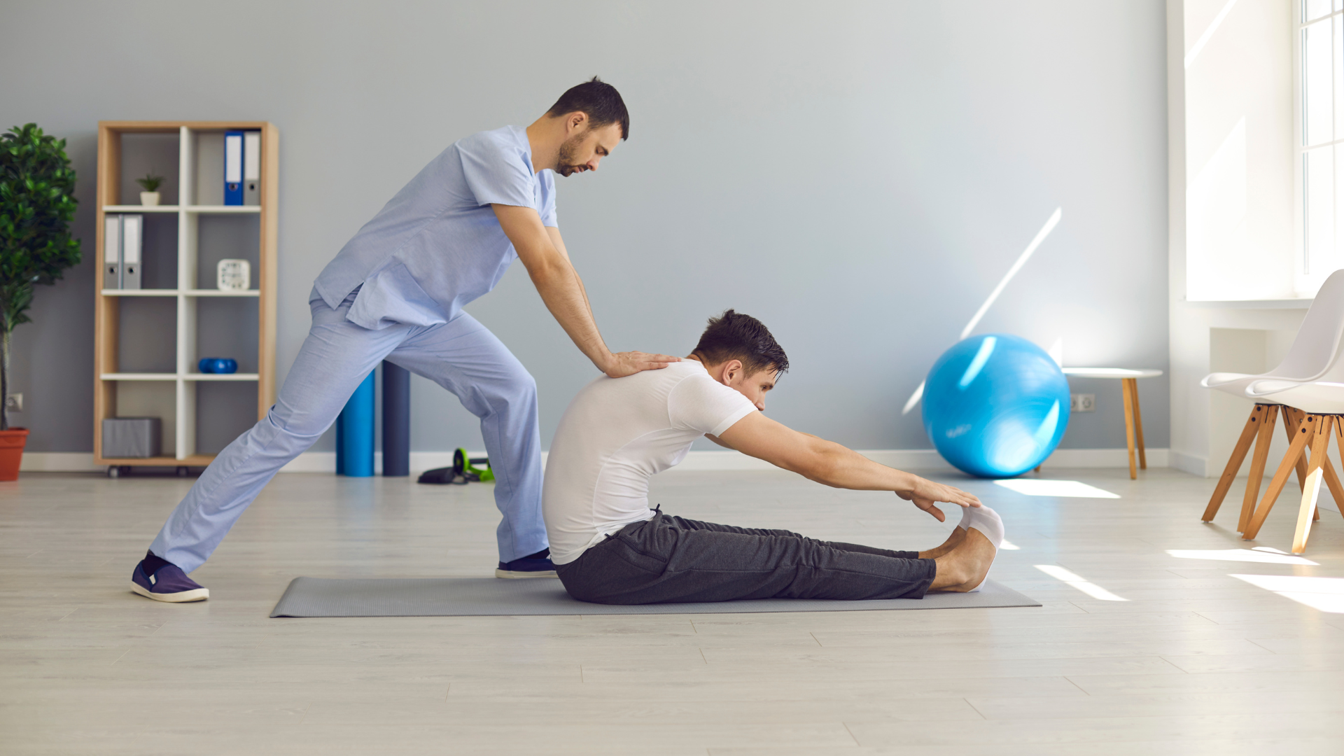 A man is sitting on a yoga mat while a nurse helps him stretch his legs.