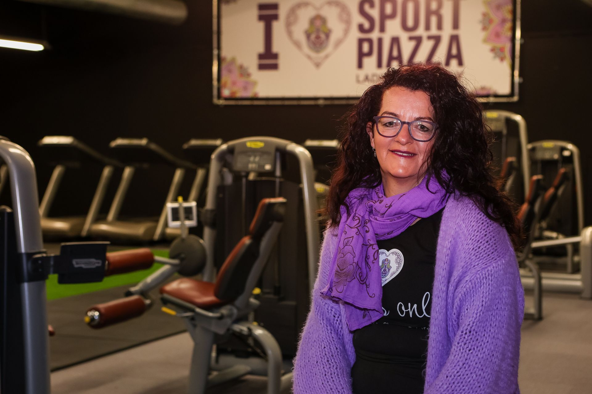 Een vrouw in een paarse trui en sjaal staat in een sportschool.