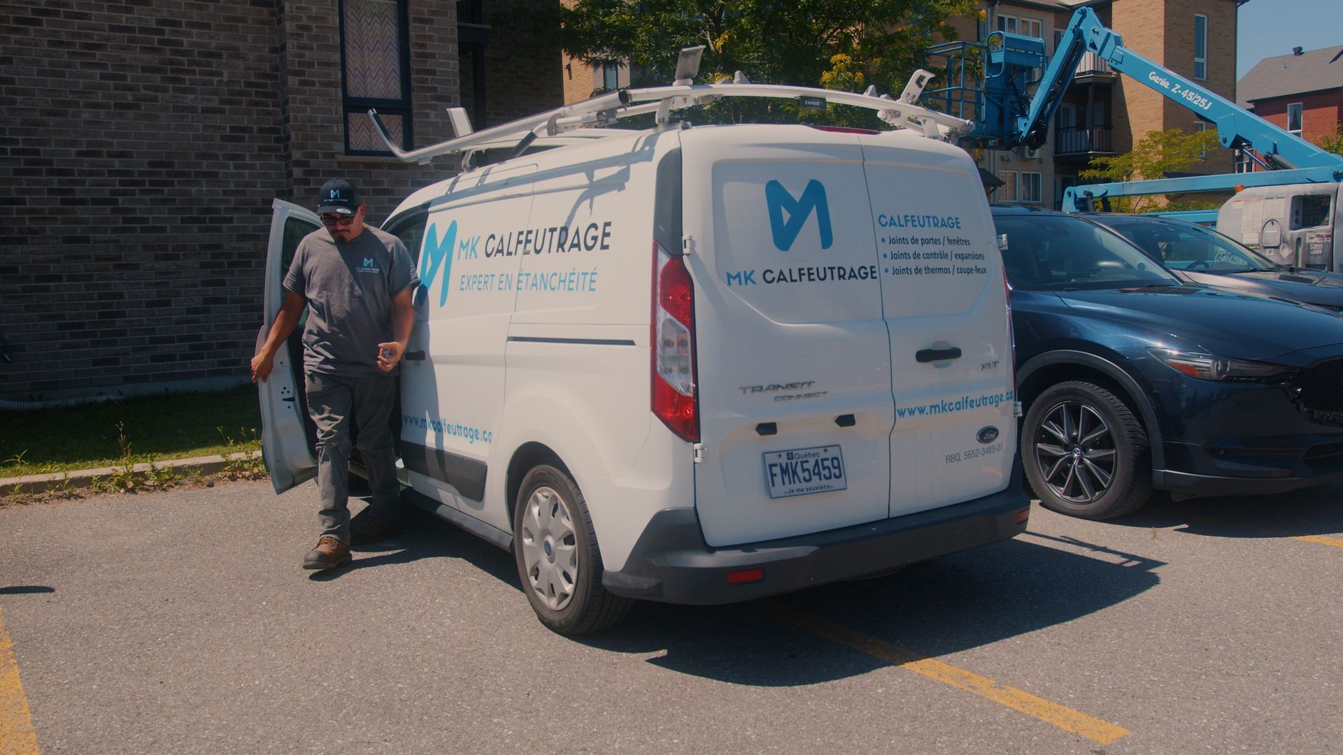 Man exiting white service van with company logo. Van parked near building and cars on sunny day.