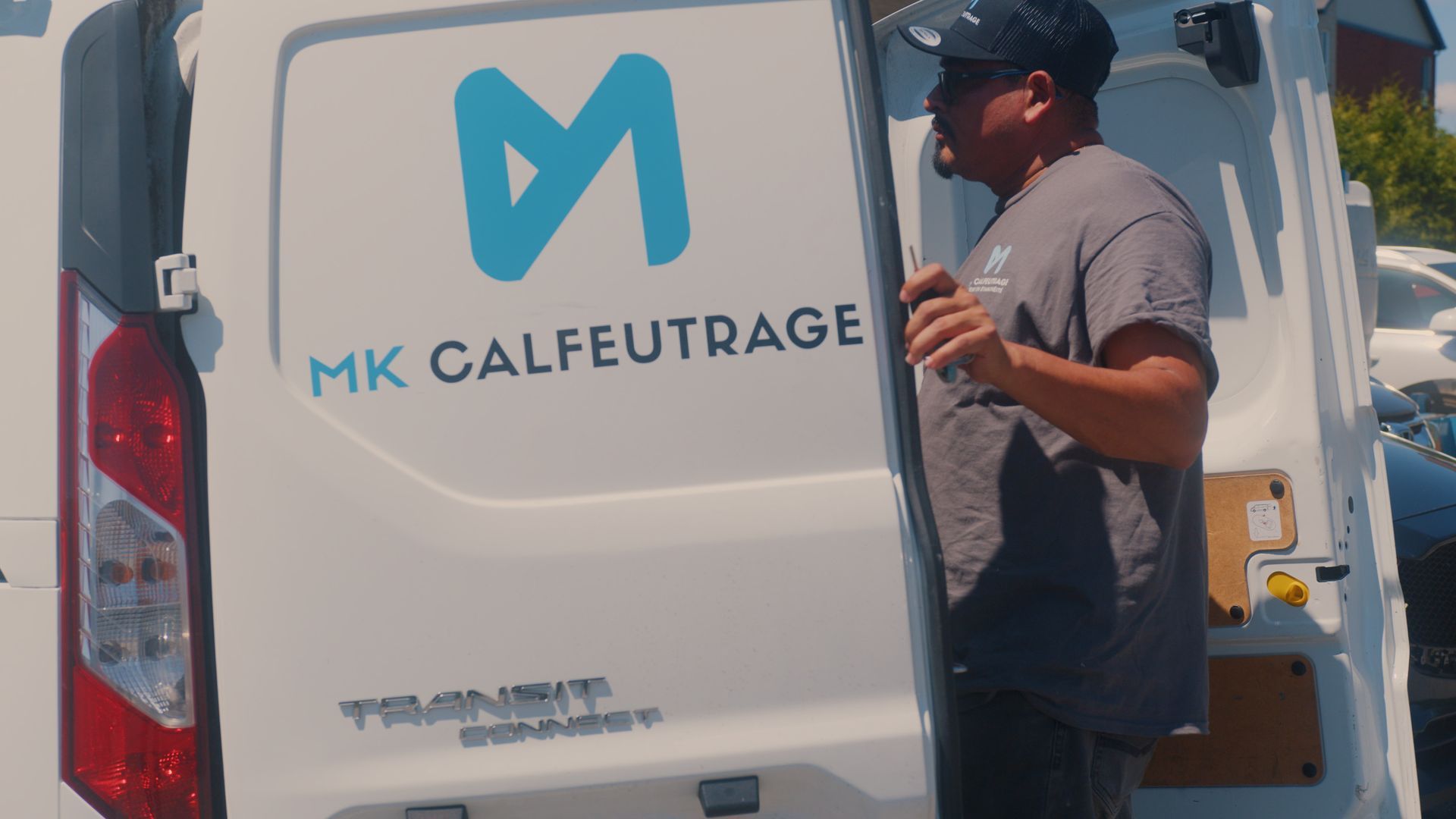 Homme près d'une camionnette blanche avec le logo « MK Calfeutrage ». Extérieur ensoleillé.