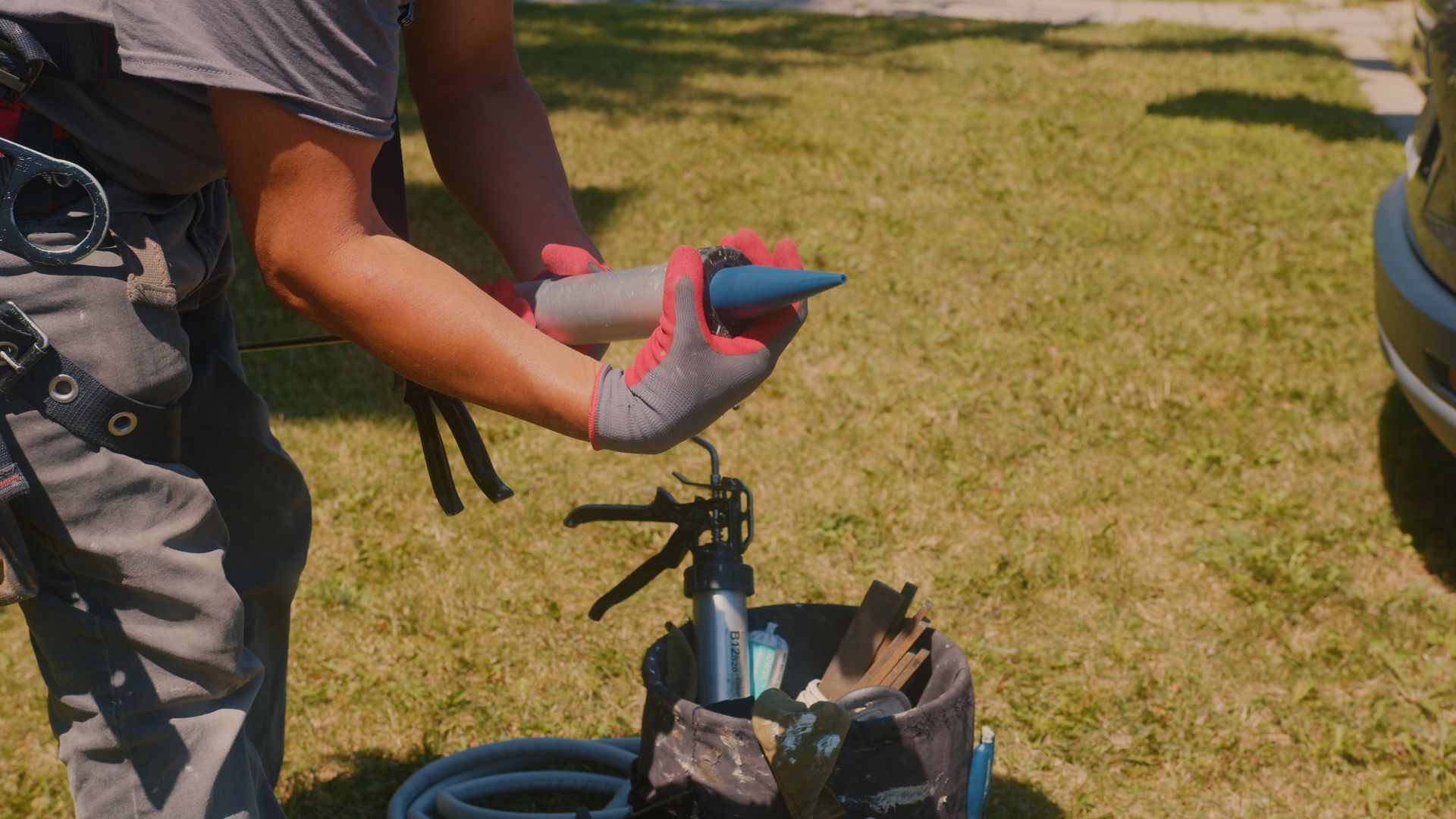 Une personne tenant un objet à pointe bleue, probablement du mastic, avec un pistolet à calfeutrer à proximité, à l'extérieur, sur une pelouse herbeuse.