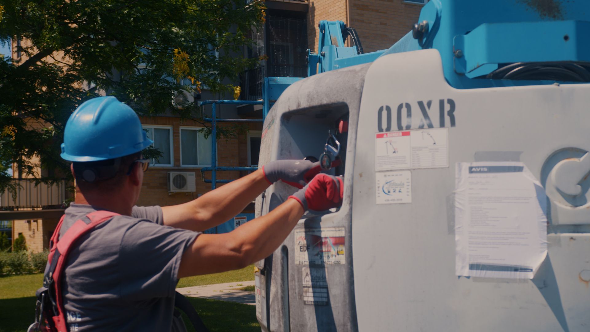 Construction worker in blue hard hat and red gloves working on a blue and gray lift machine outside a building.