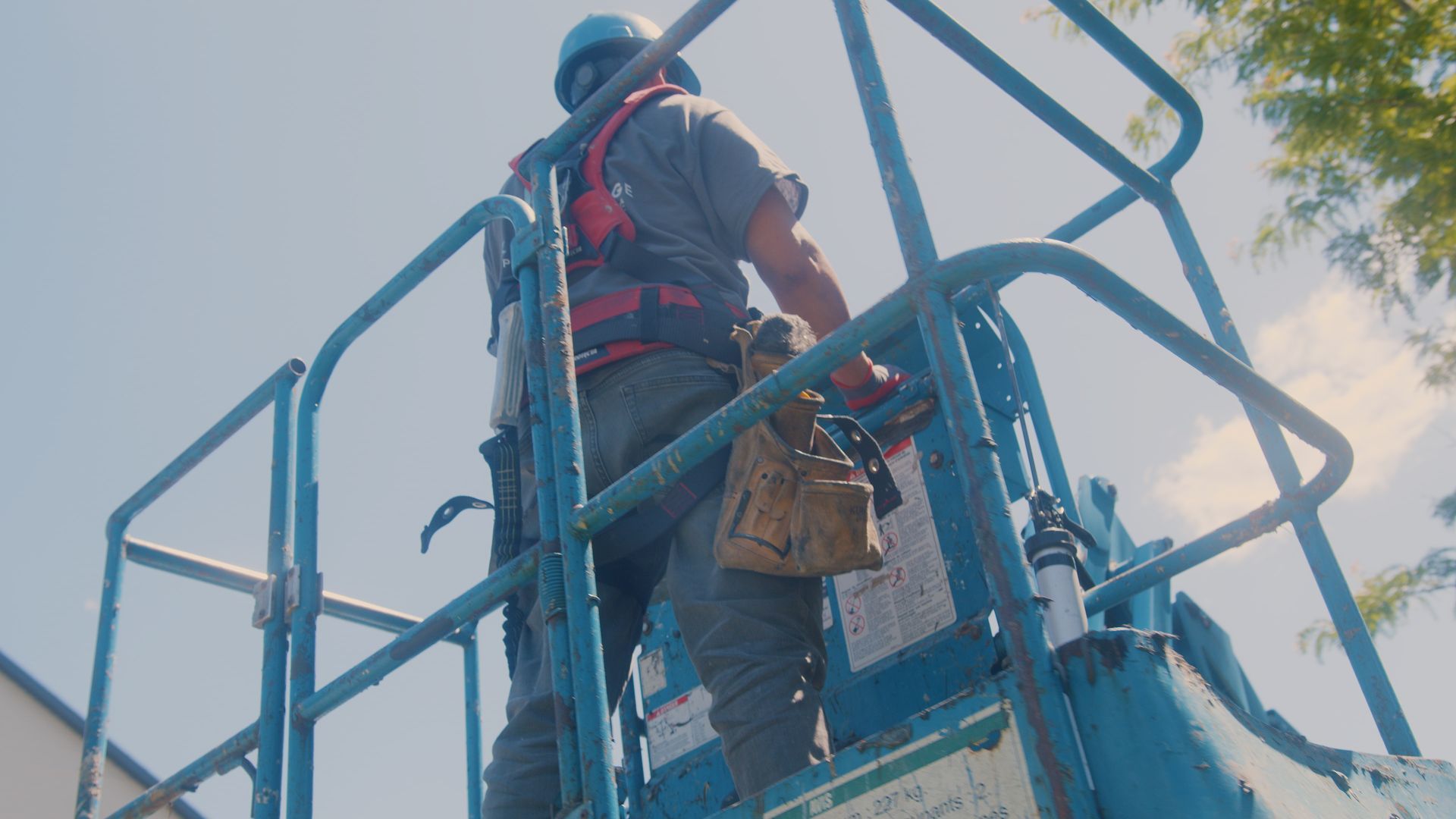 Un homme en tenue de sécurité travaille sur une plateforme élévatrice bleue, à l'extérieur d'un bâtiment. Ciel bleu en arrière-plan.