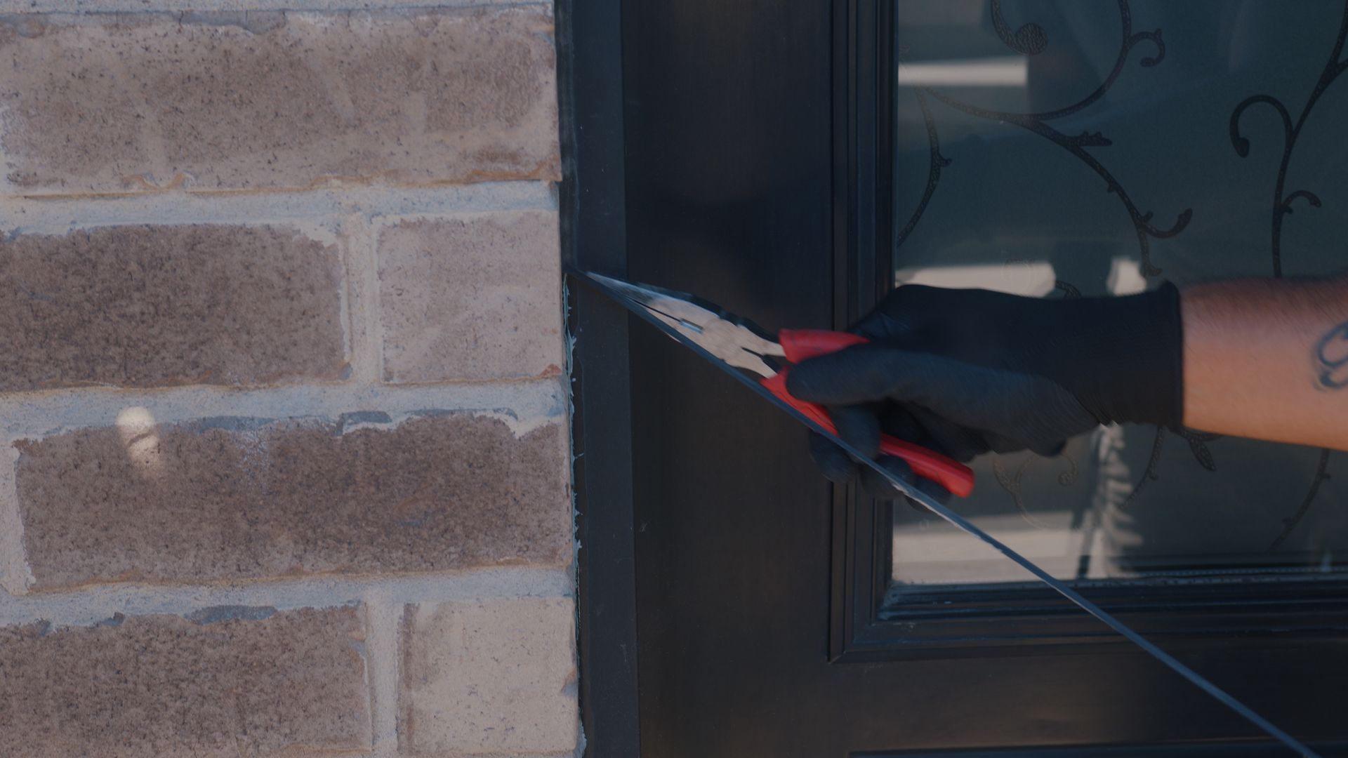 Main gantée tenant un coupe-fil contre un cadre de porte noir, à côté d'un mur de briques.