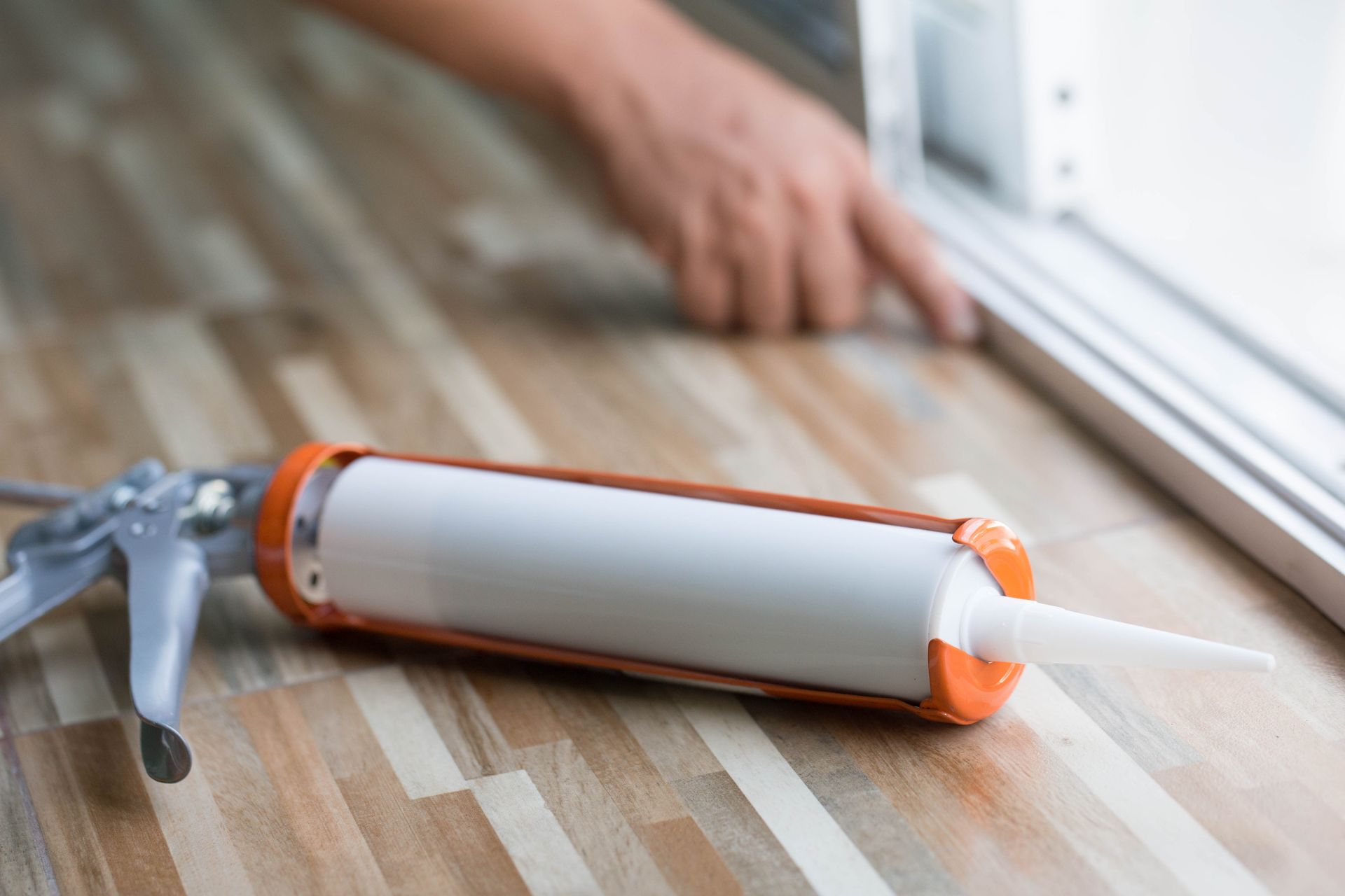Caulk gun and tube on floor, person applying caulk to window frame.