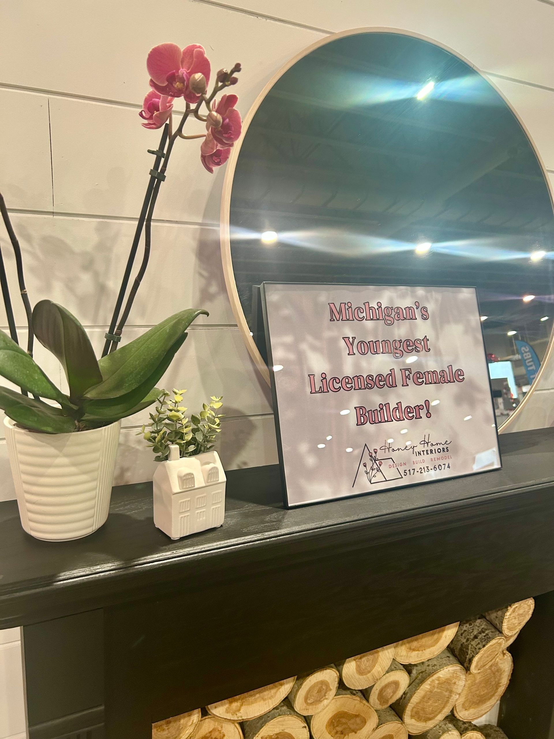 Orchid, small plant, and sign displayed on a mantel with a mirror and stacked logs.