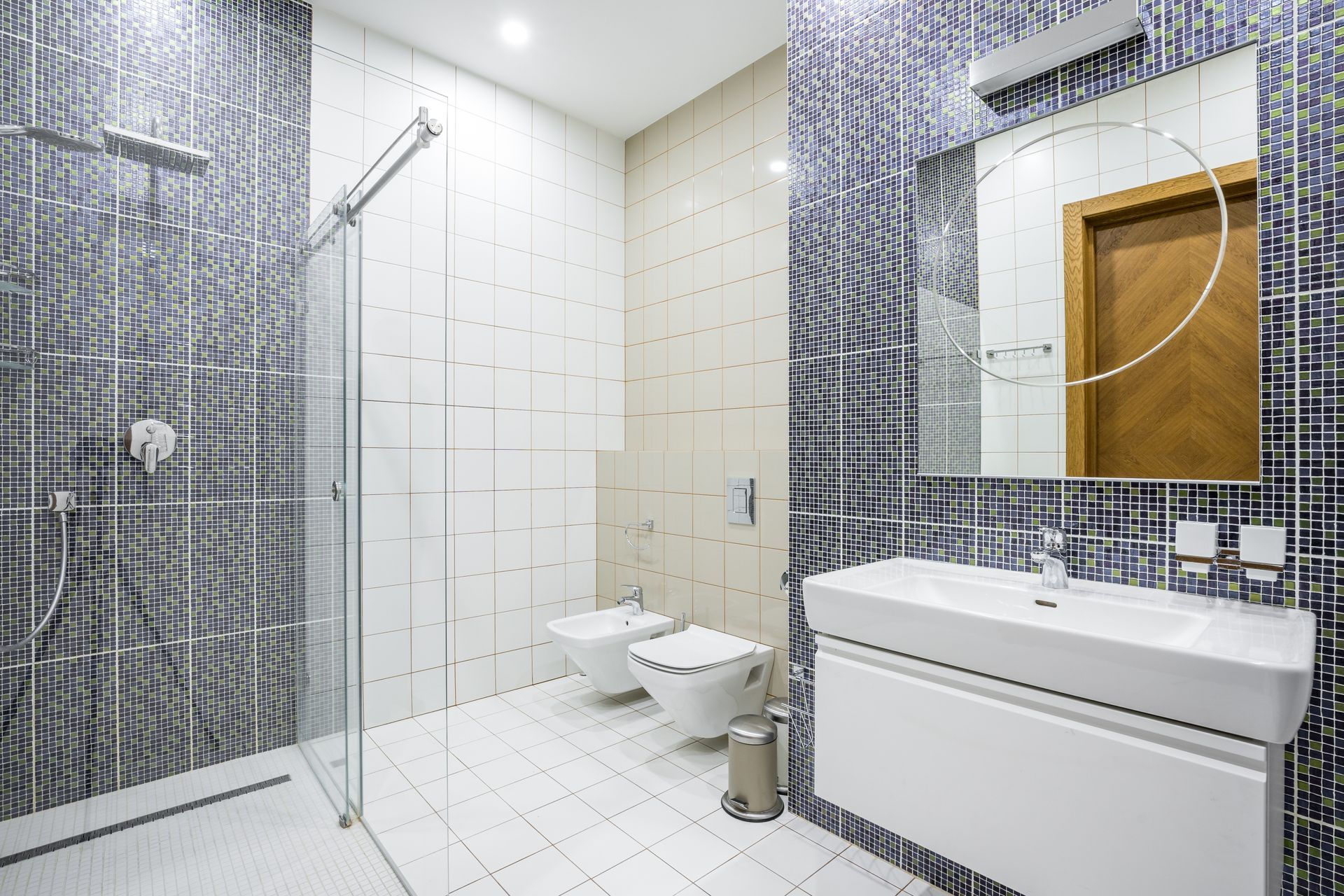 A modern bathroom with a glass shower, white bidet and toilet, and a white vanity with a mirror against patterned tile.