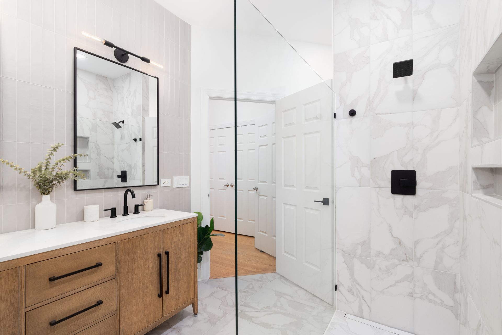 Modern bathroom with a wooden vanity, marble shower, and black fixtures in a bright, neutral space.