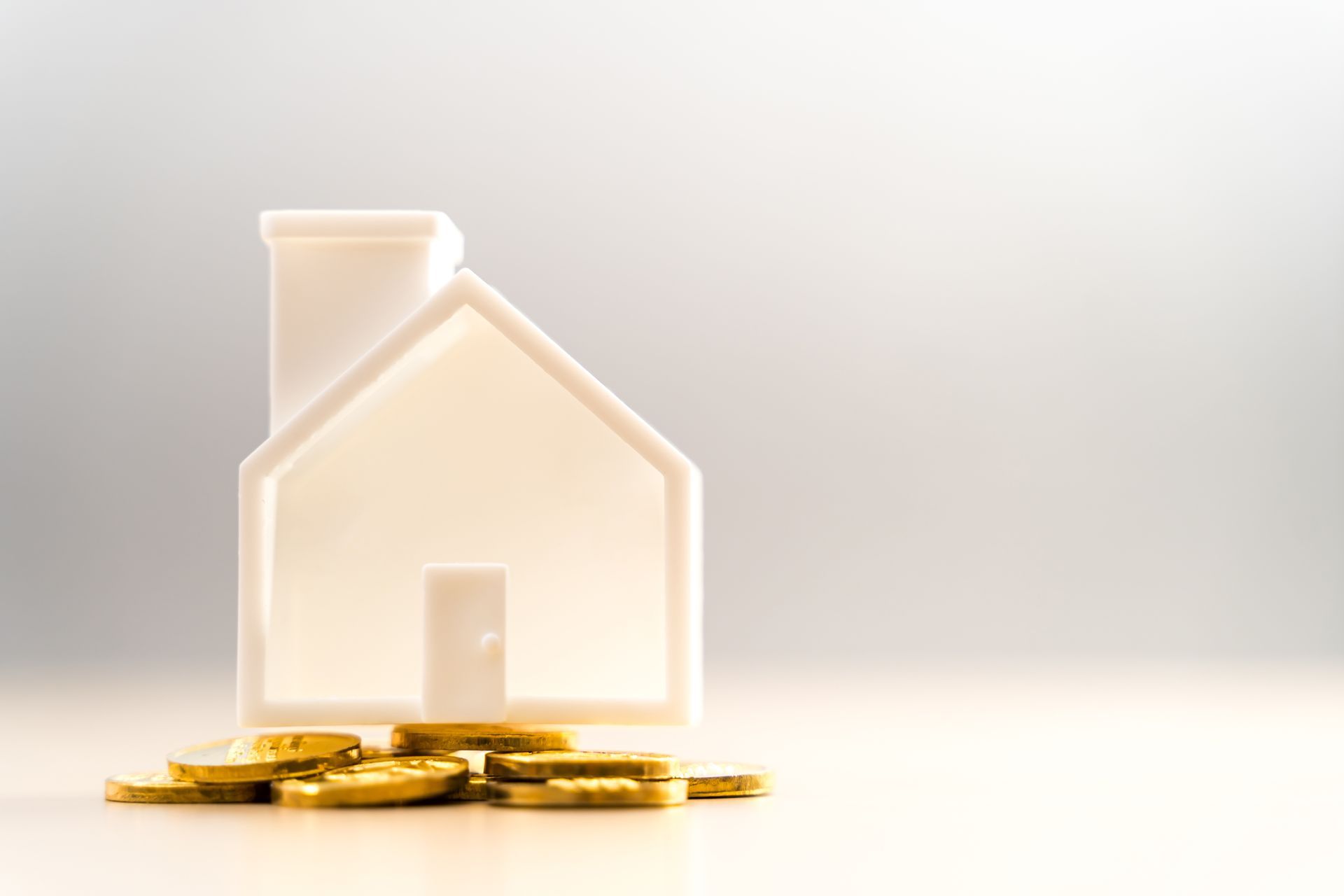 A white 3D house model standing on a small pile of gold coins against a soft, light-colored background.