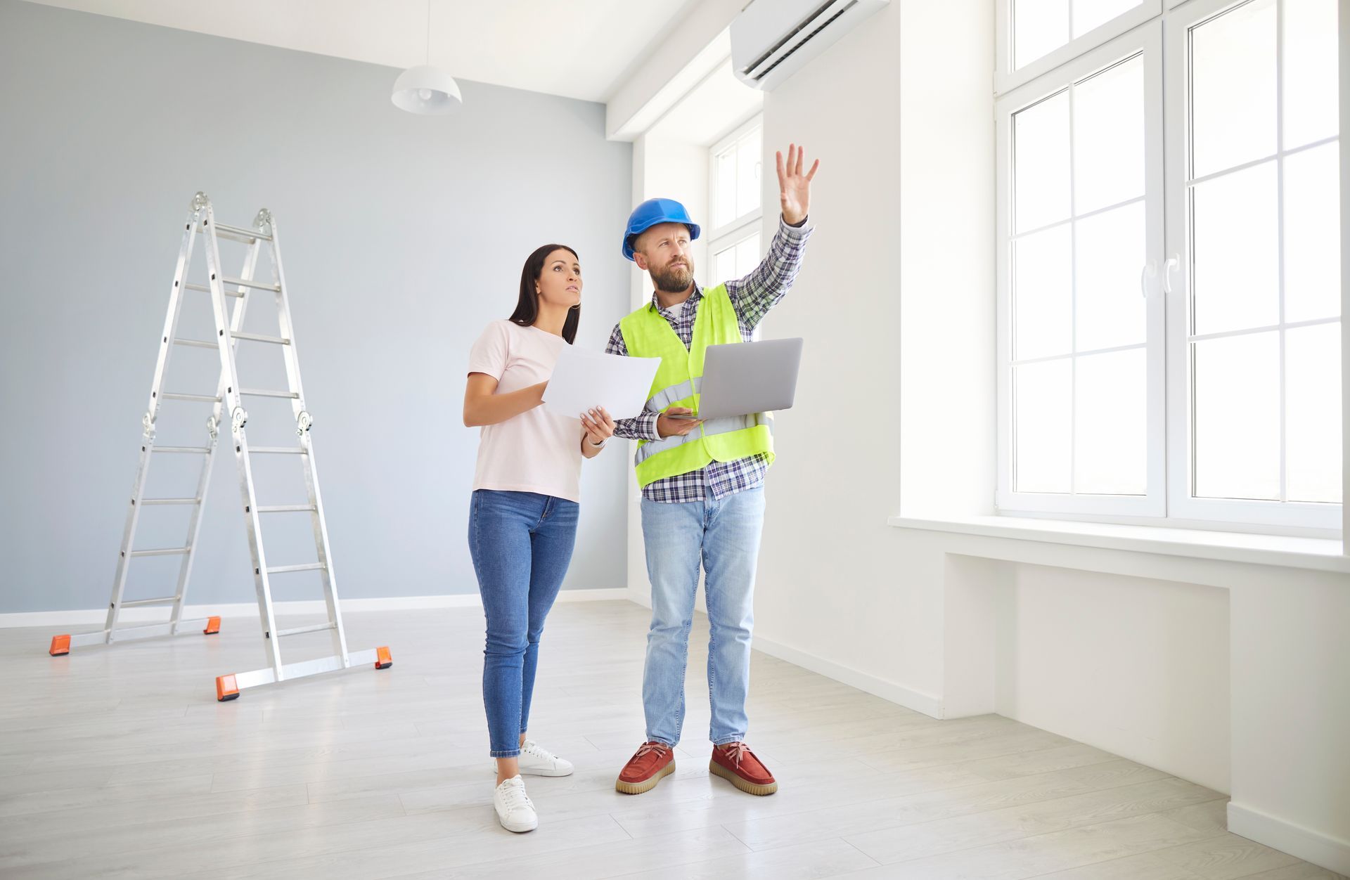 A construction professional in a hard hat and safety vest points at a window while reviewing plans with a client indoors.