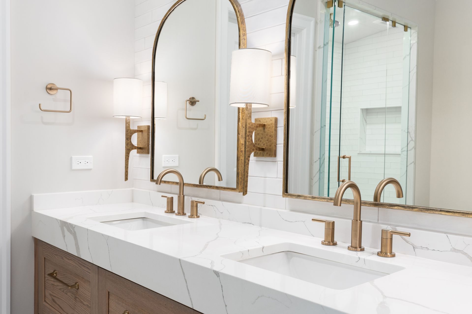 A modern bathroom vanity with dual sinks, gold fixtures, arched mirrors, and marble countertops against white subway tile.