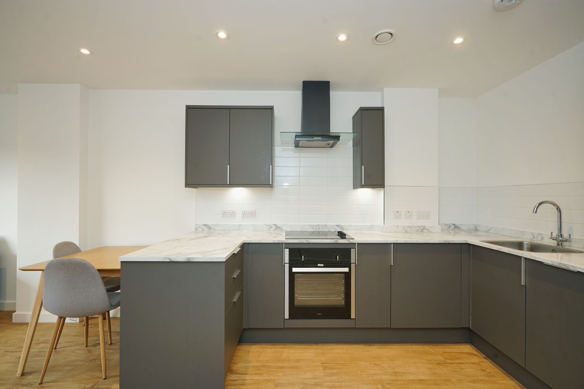 Modern kitchen with grey cabinetry, white marble-style countertops, built-in oven, and a wooden dining table with chairs.