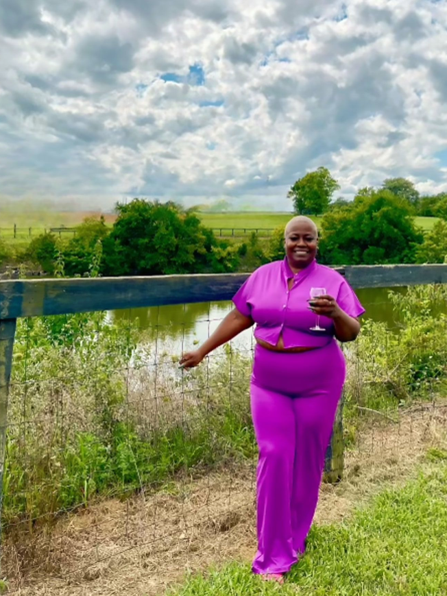 Woman in purple outfit holding wine, smiling outdoors near a pond and fence.