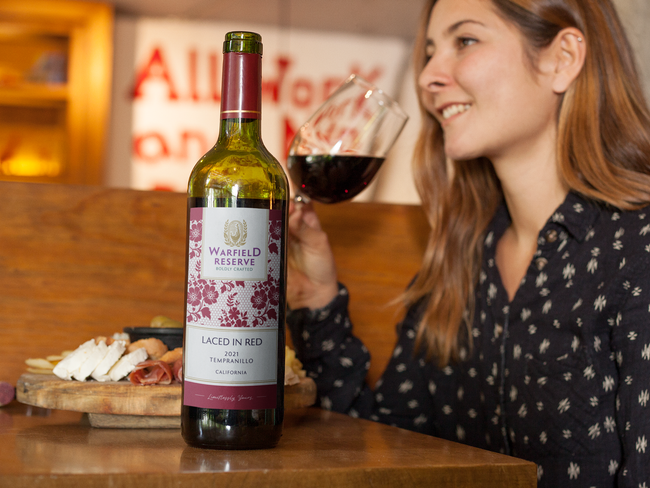 Woman smelling red wine, bottle of wine on table, wooden setting.