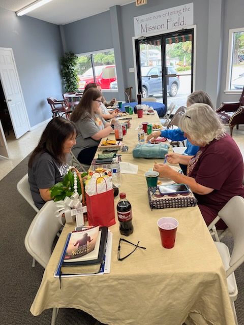 People seated around a table crafting. Supplies, drinks, and a small gift bag are present. Window view in background.