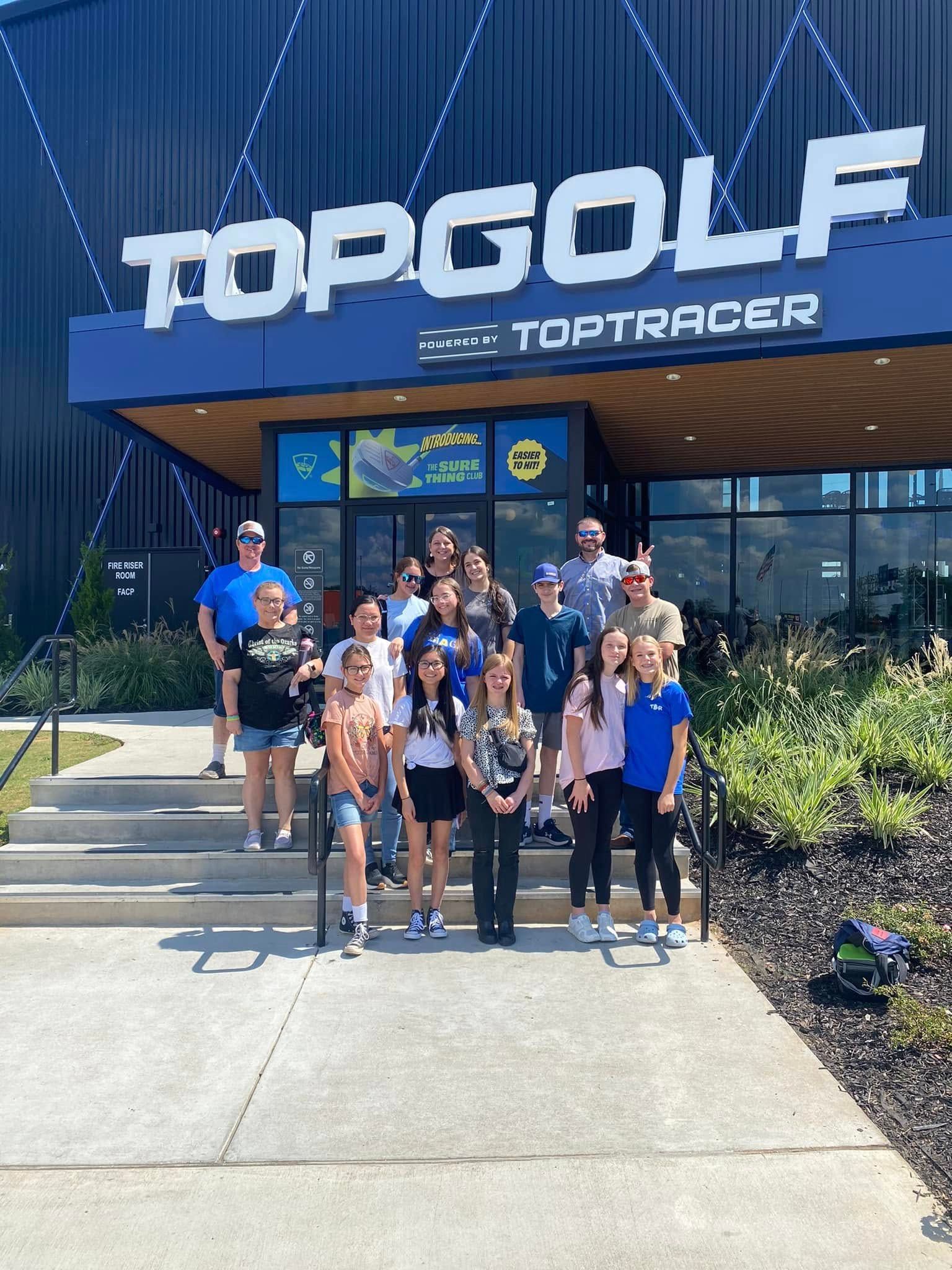 Group of people standing in front of Topgolf building, blue and white sign, sunny day.