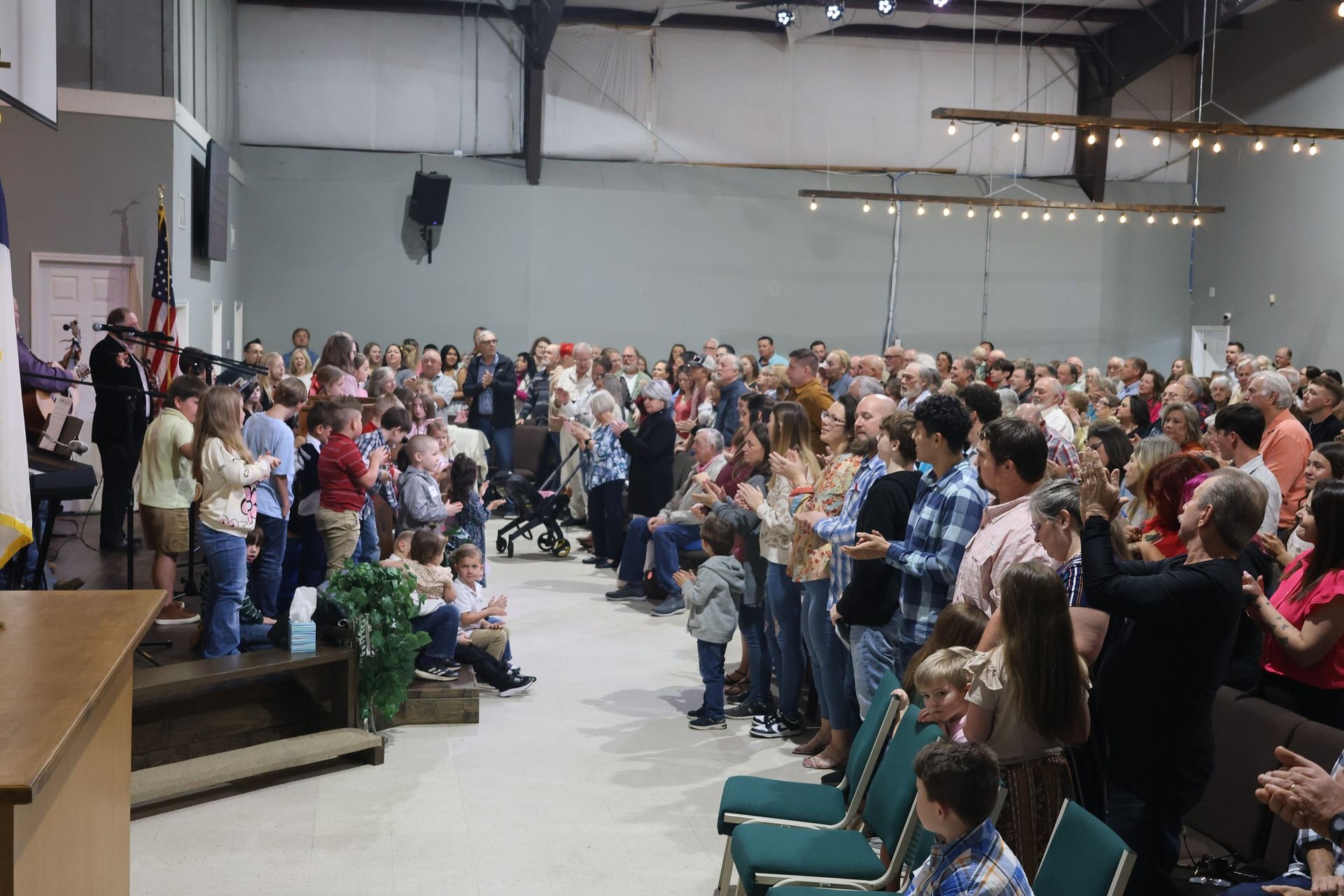 Large crowd applauding in a brightly lit room. A few people stand near a podium.