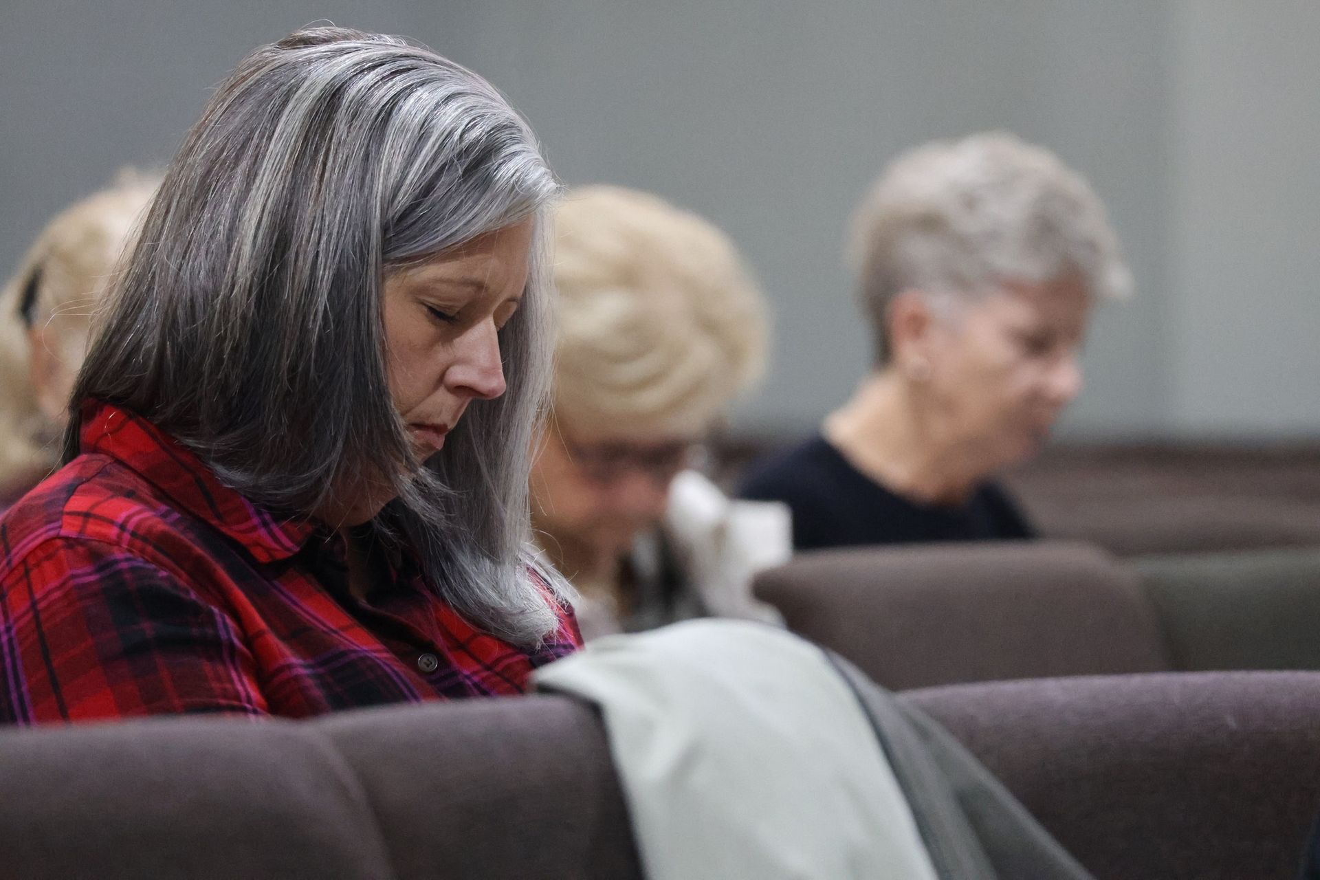 Three people with bowed heads sit in a row of pews, eyes closed, possibly in prayer.