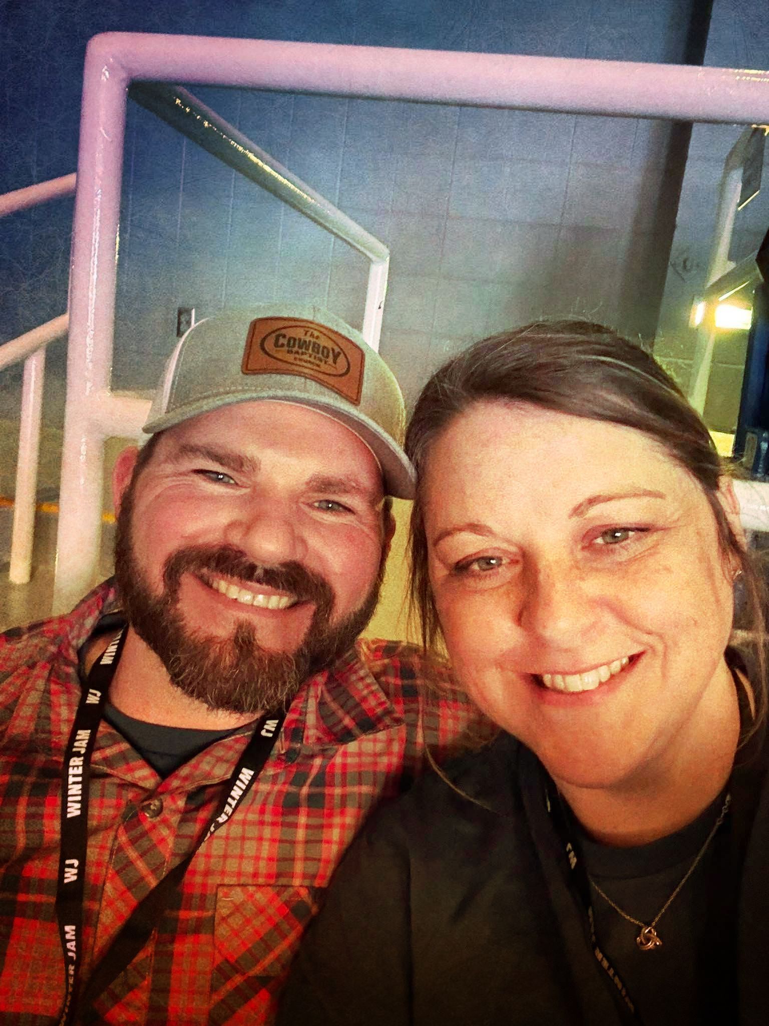 Smiling couple, man in plaid shirt and cap, woman in dark shirt, indoors.
